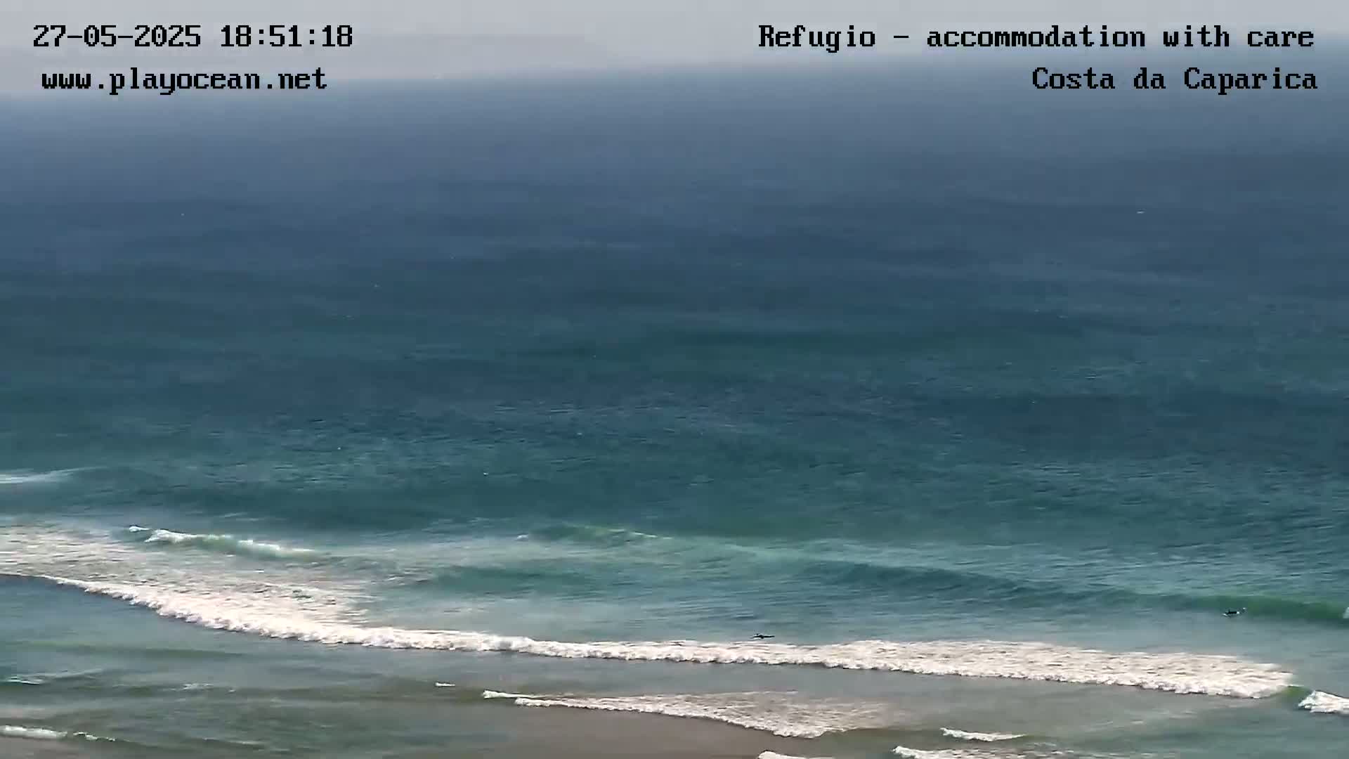 An overcast sky hangs over a teal ocean with several surfers riding waves that are breaking near a sandy shore.