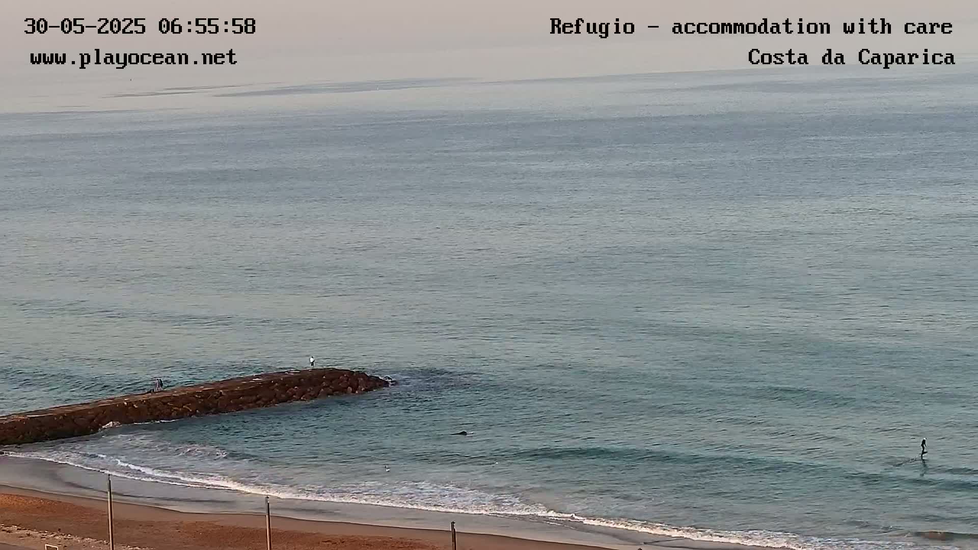 A calm, overcast day shows a sandy beach with a low rock jetty extending into a gently waving ocean.