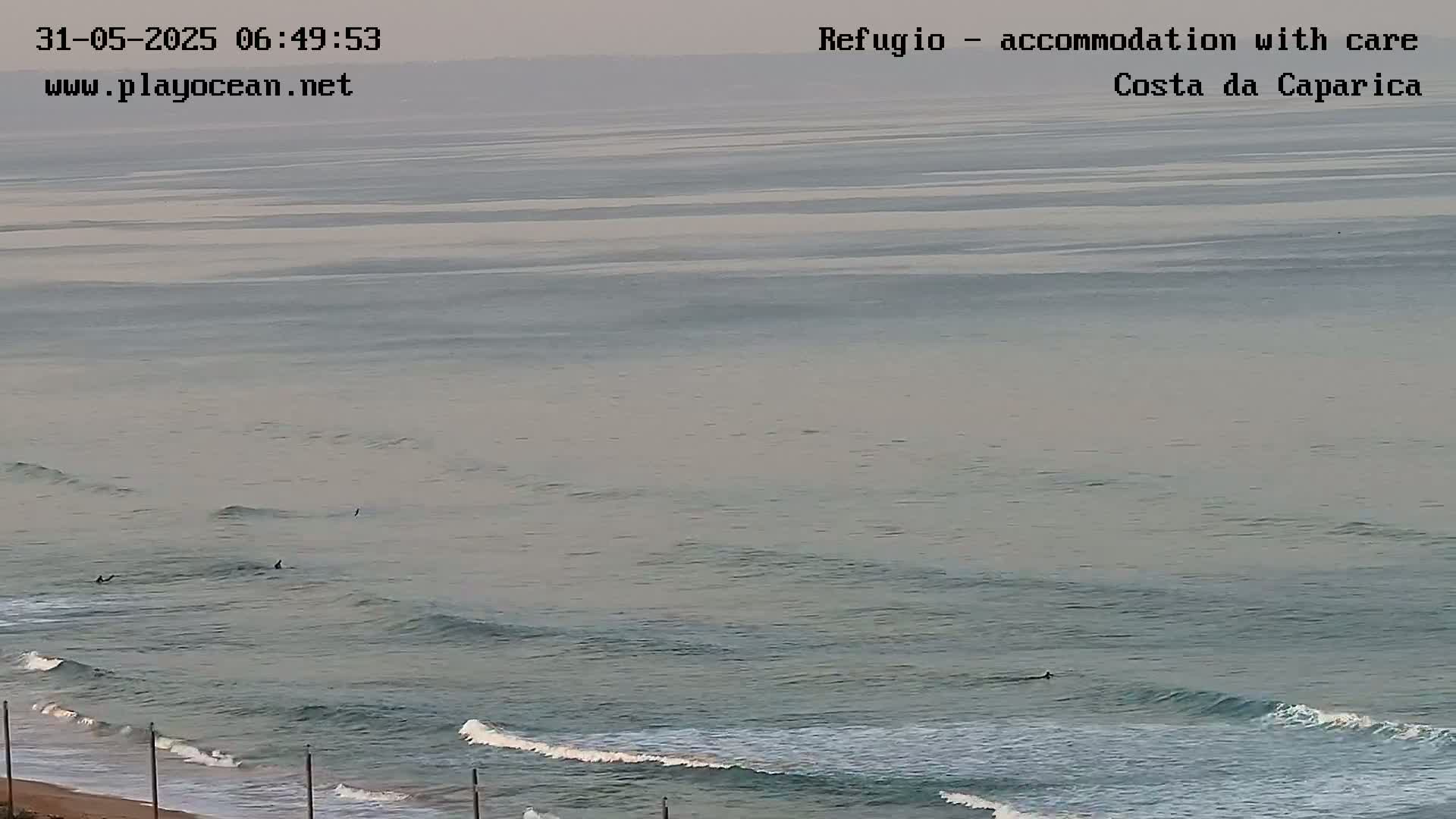 A hazy, overcast day reveals a calm ocean with small waves rolling onto a sandy beach where several surfers are visible in the water.