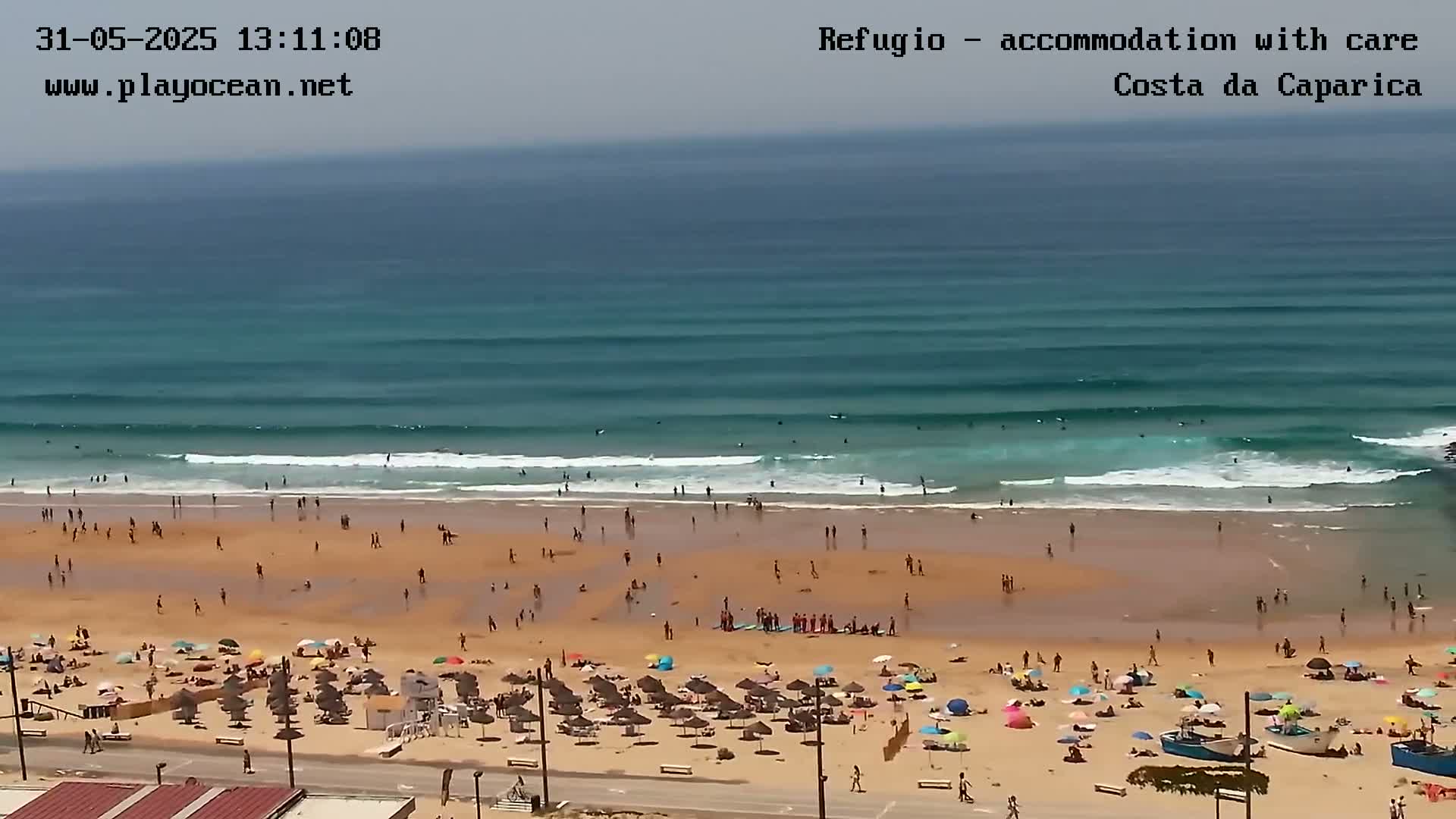 A sandy beach with many people and umbrellas is shown, with a calm ocean and gently rolling waves visible in the background under a hazy sky.