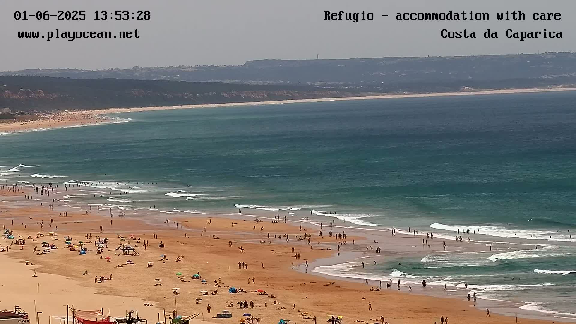 A wide shot of a sunny beach with many people on it, with teal-colored ocean waves breaking on the shore.