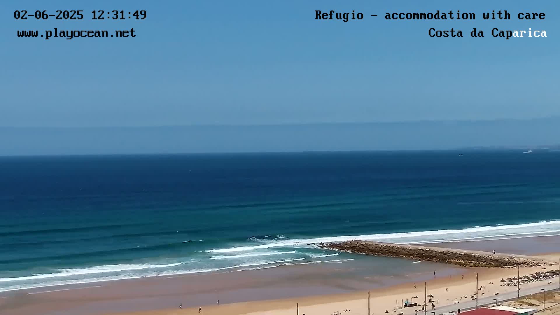 An ocean beach with small waves under a clear blue sky shows people relaxing on the sand near a rock jetty.