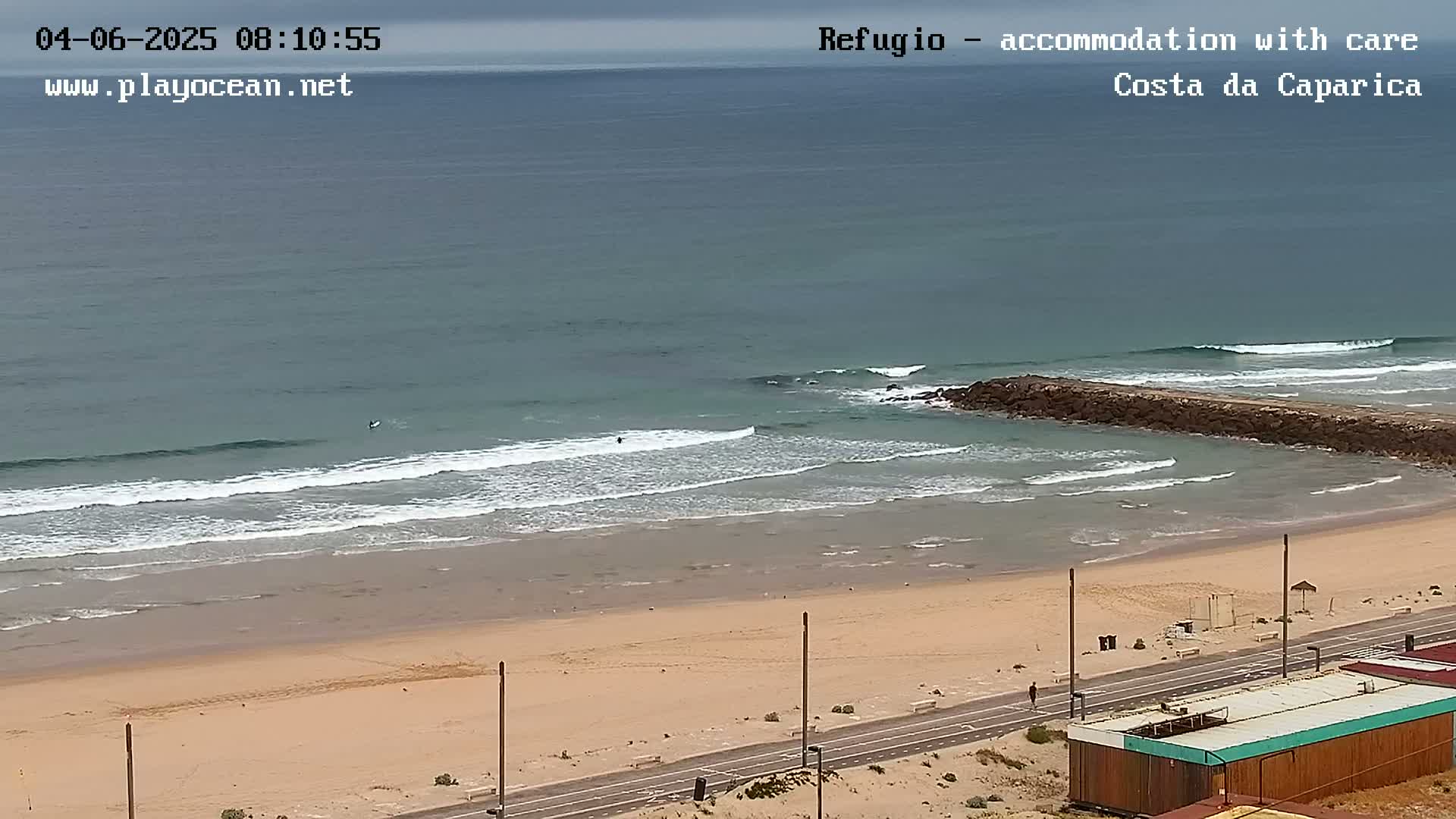 An overcast day shows a sandy beach with small waves lapping the shore, a road nearby, and a few surfers in the water.