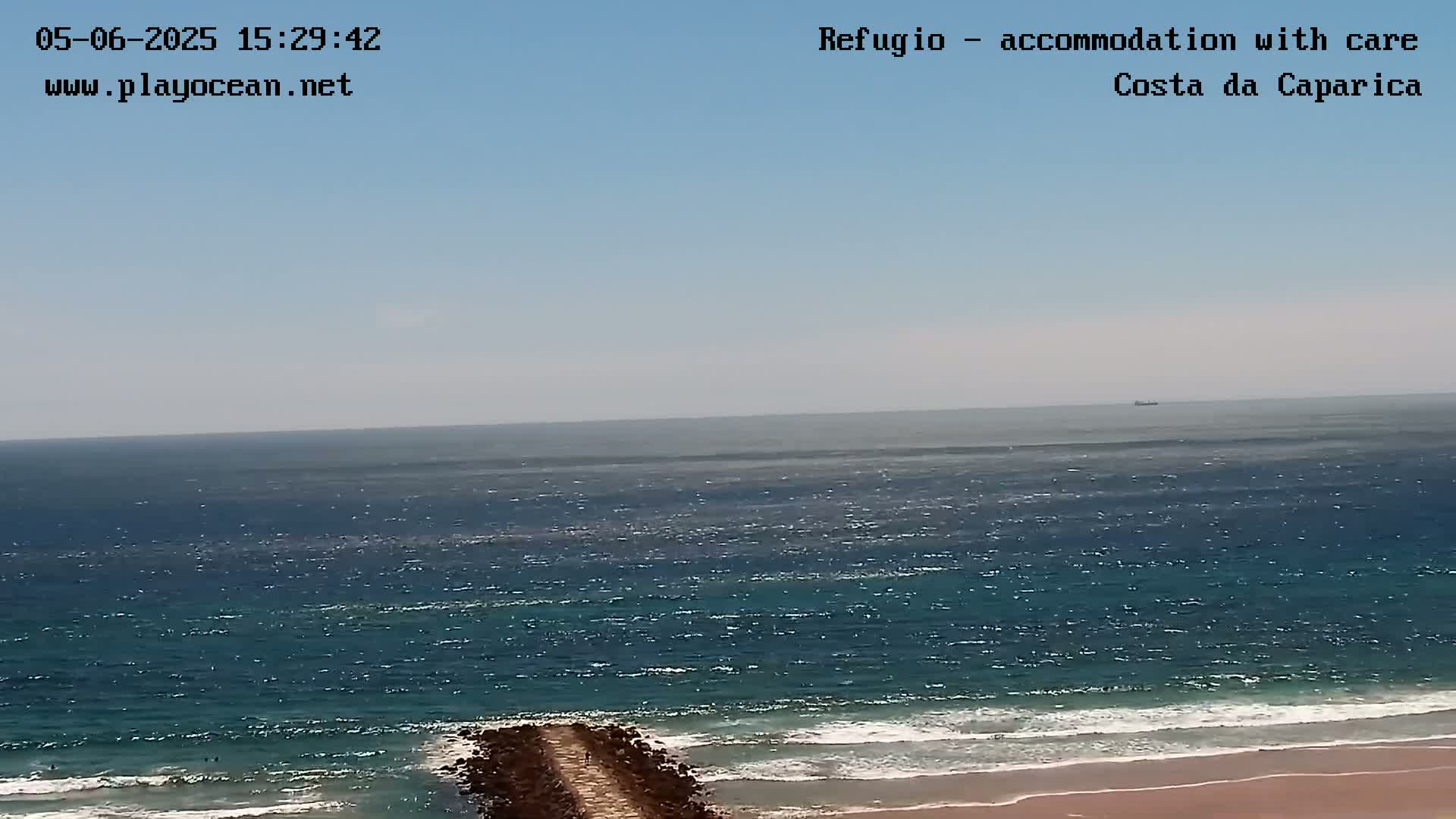 A mostly clear sky overlooks a wavy ocean meeting a sandy beach with a dark rock jetty extending from the shore.