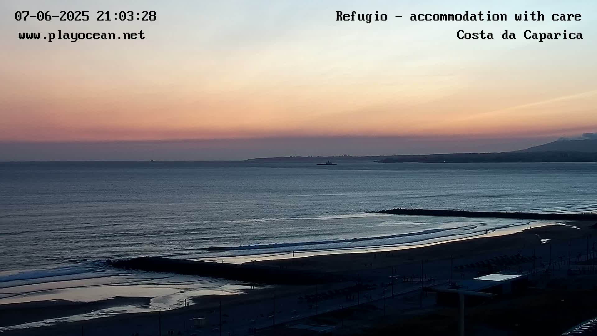 A tranquil ocean beach scene at sunset, featuring calm waves rolling onto the shore and a distant mountain range visible on the horizon.