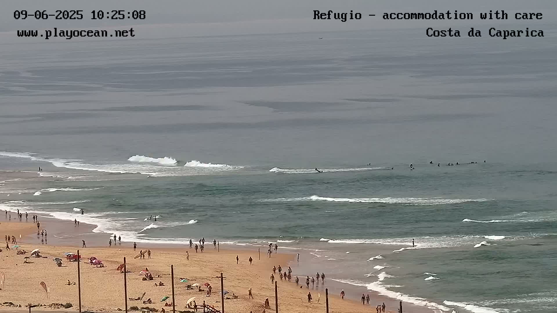 A sandy beach with numerous people on it is partially visible in the foreground, leading to an ocean with moderate waves and several surfers visible in the distance under an overcast sky.
