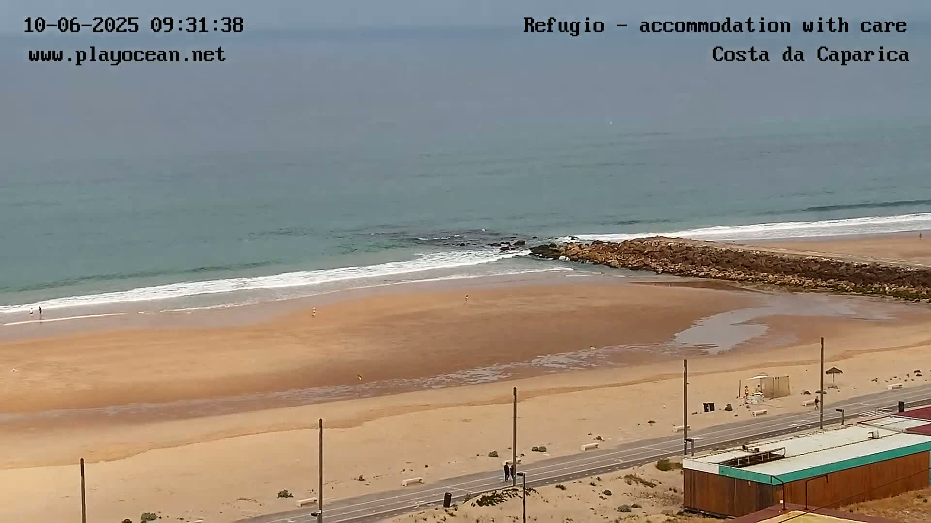 A sandy beach with a calm ocean, a rocky jetty, and a paved walkway next to some small buildings under an overcast sky.