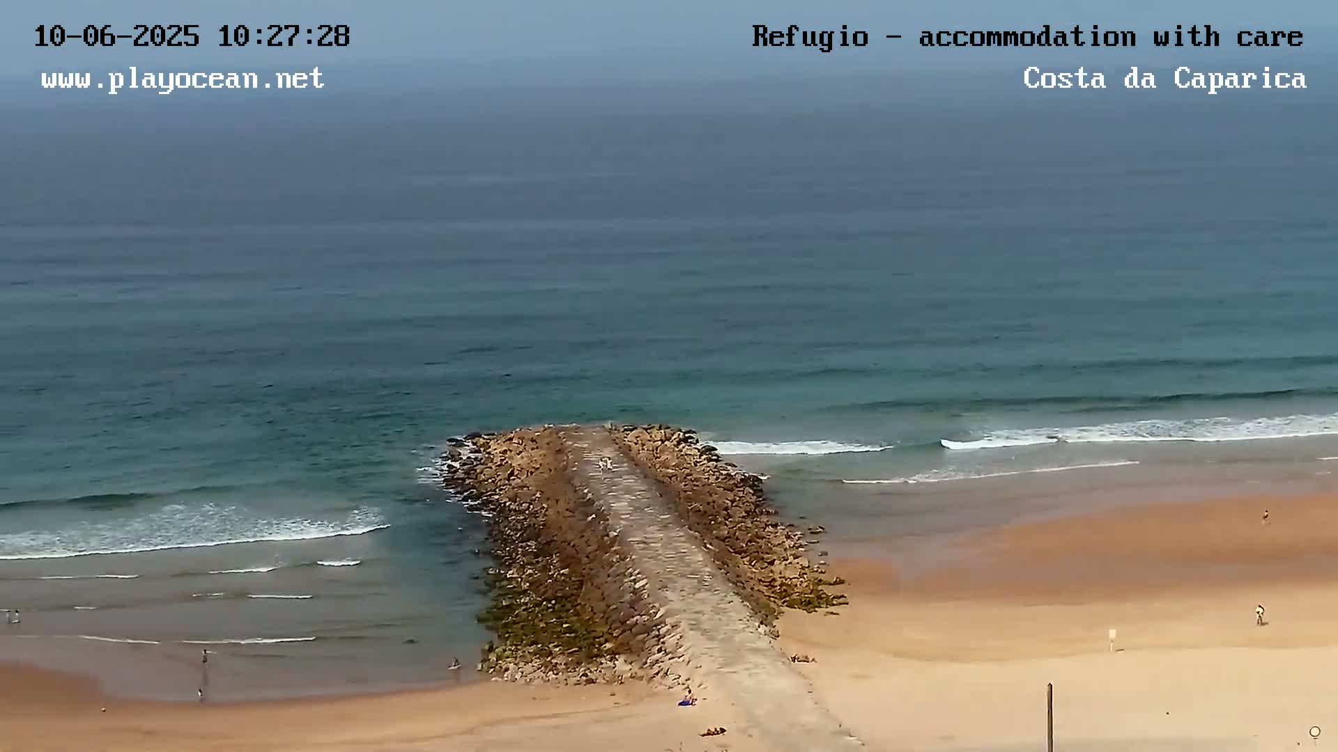 An aerial view shows a rocky jetty extending from a sandy beach into an ocean with small waves under an overcast sky.