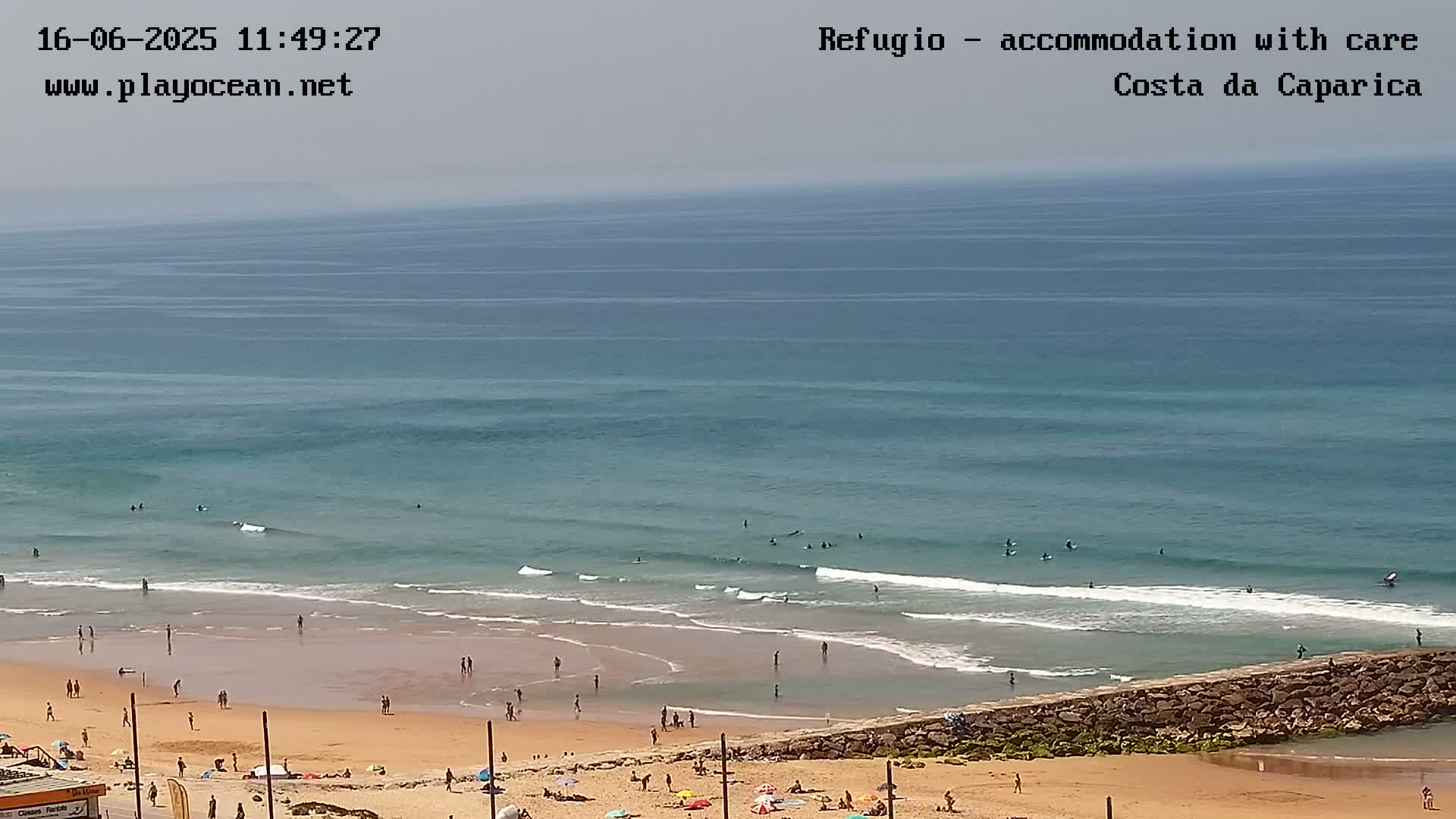 A sandy beach with people sunbathing and swimming, next to a calm ocean with several surfers, under a mostly clear sky.