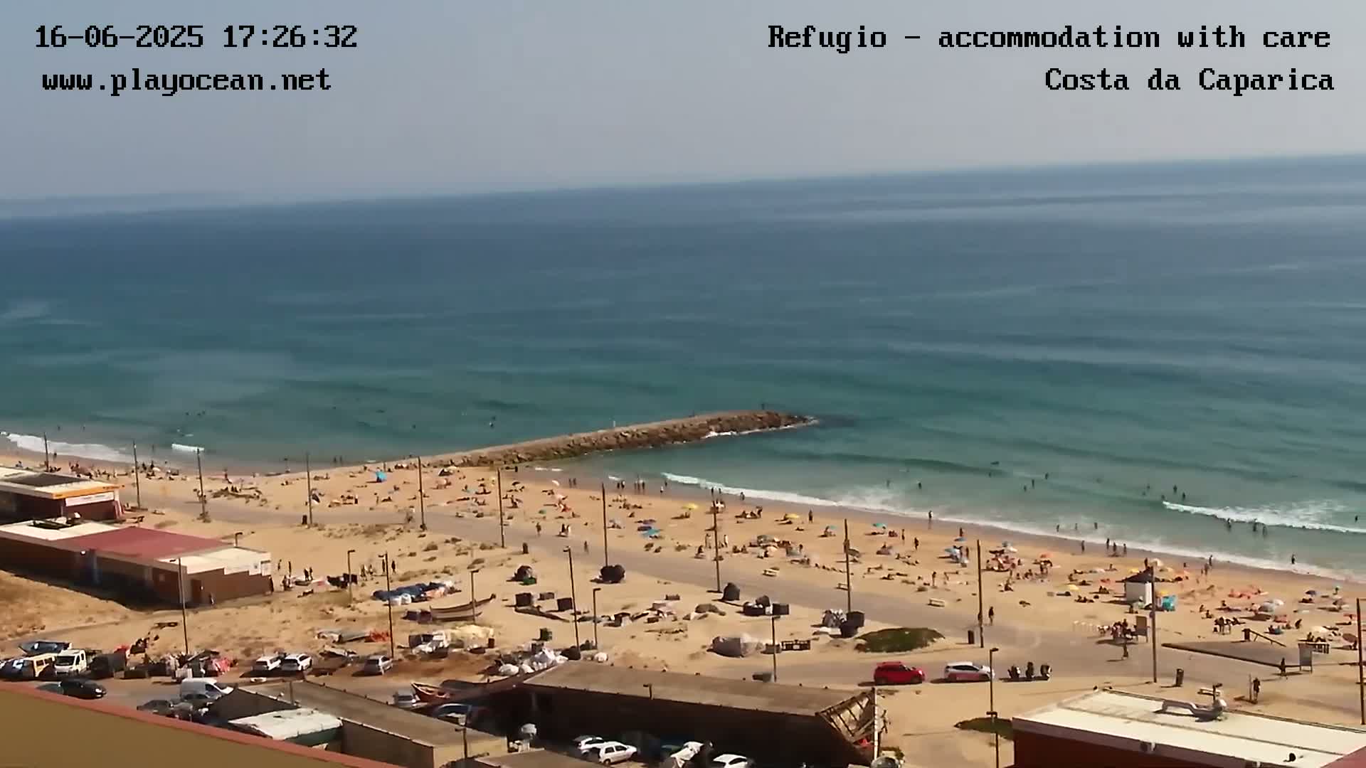 A sunny day at a beach shows many people sunbathing and swimming in the ocean, with a parking lot and buildings visible nearby.