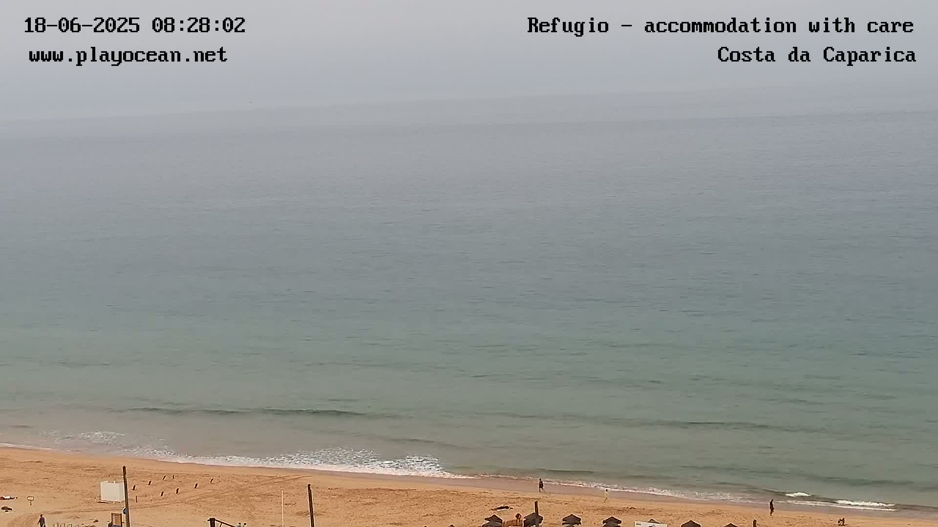A hazy, overcast day reveals a sandy beach with calm, light-green ocean waves gently lapping the shore.