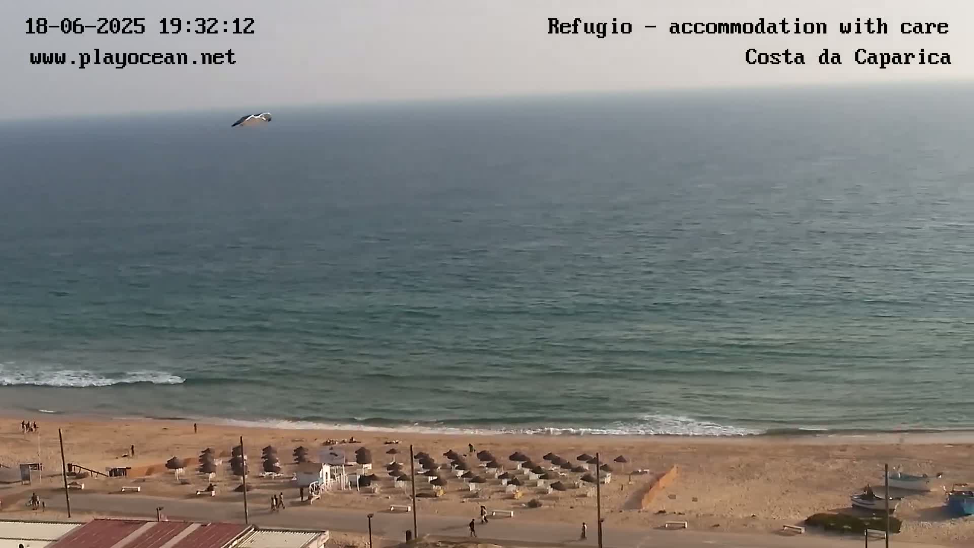 A mostly clear day shows a calm ocean with small waves lapping a sandy beach lined with beach umbrellas, a few people, and some boats.