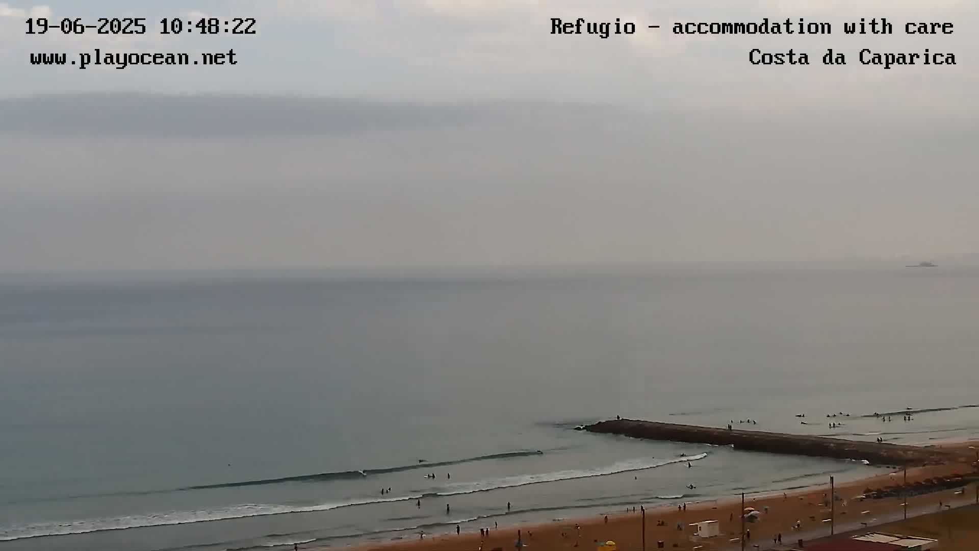 A hazy, overcast day reveals a beach scene with people swimming and surfing near a breakwater, extending into a calm ocean.