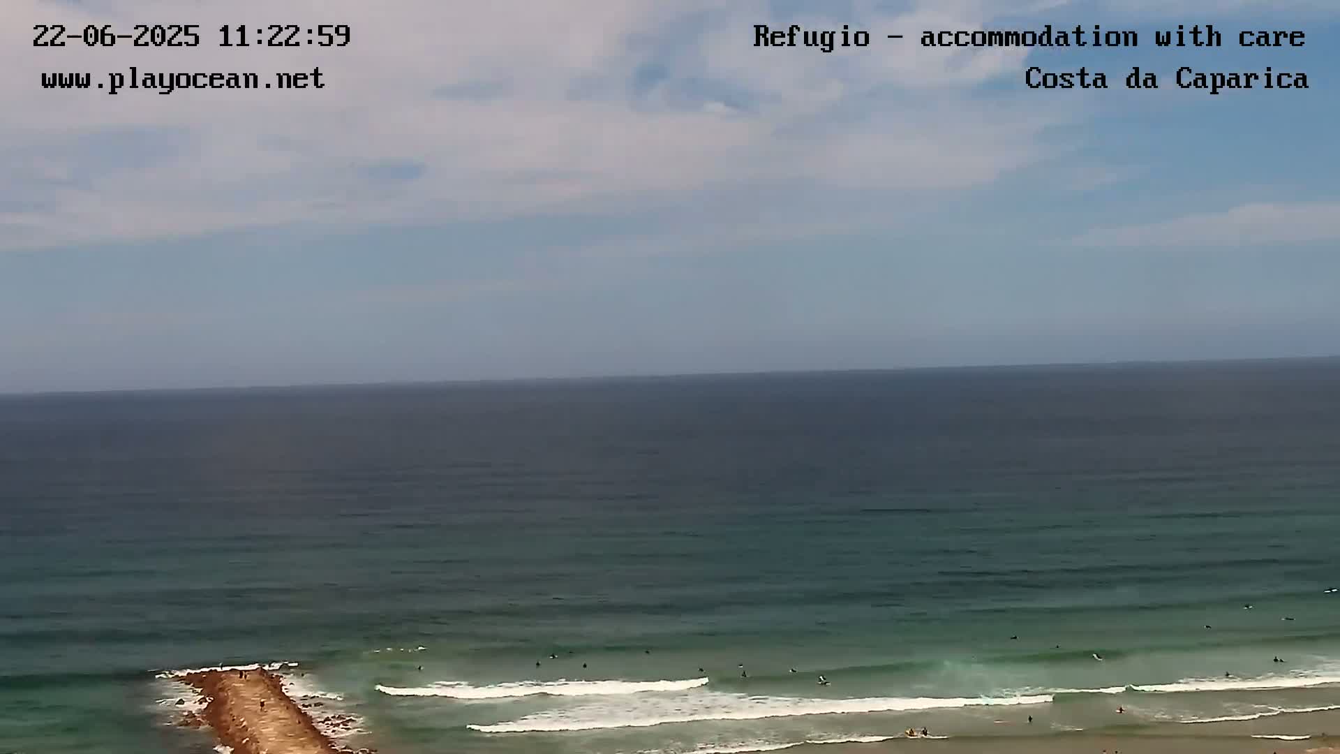A long shot of an ocean under a partly cloudy sky shows surfers riding waves near a rocky jetty.