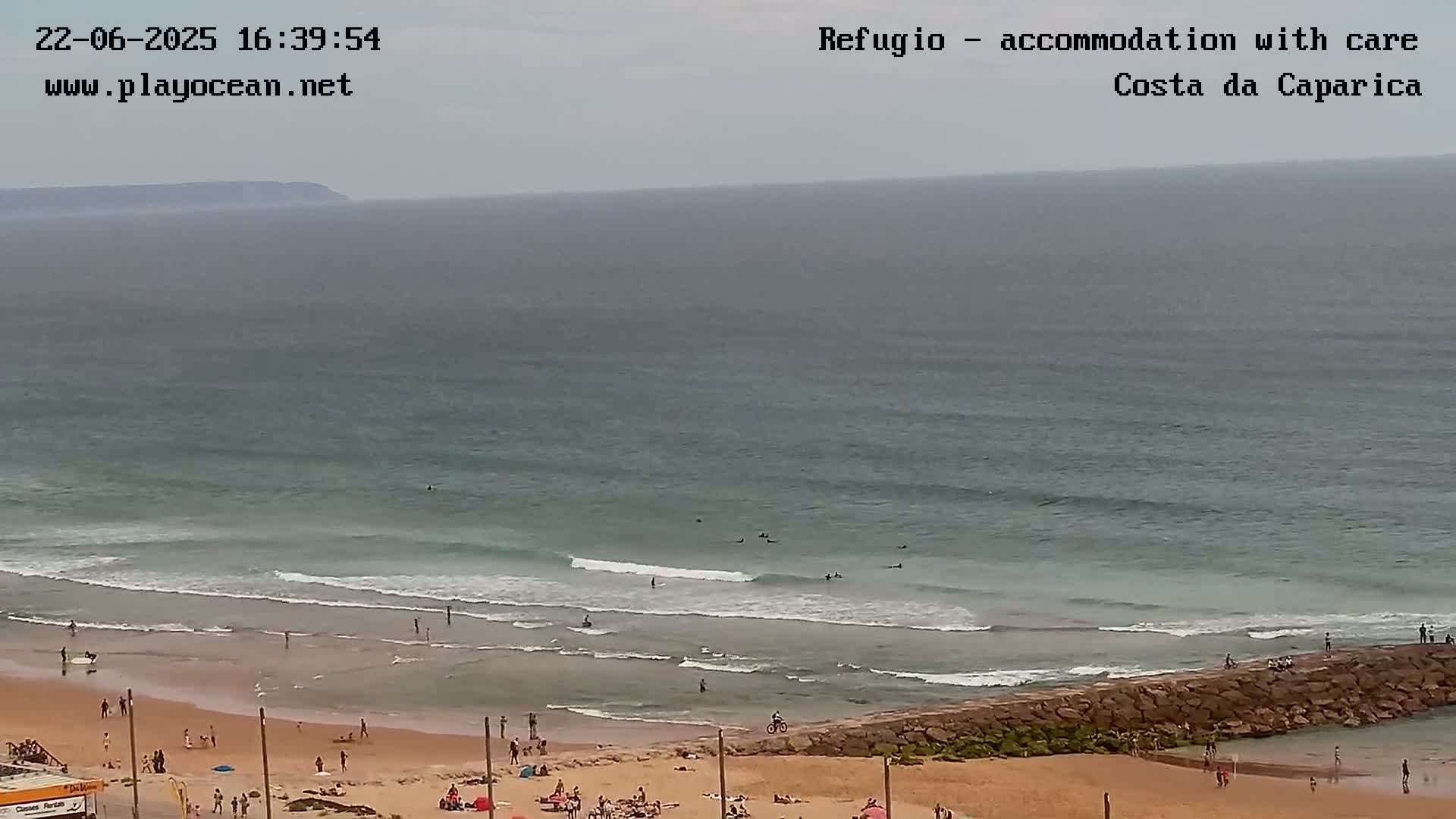 A beach scene on an overcast day shows a sandy shore with people sunbathing and swimming, small waves rolling onto the sand, and a distant landmass visible in the hazy ocean.