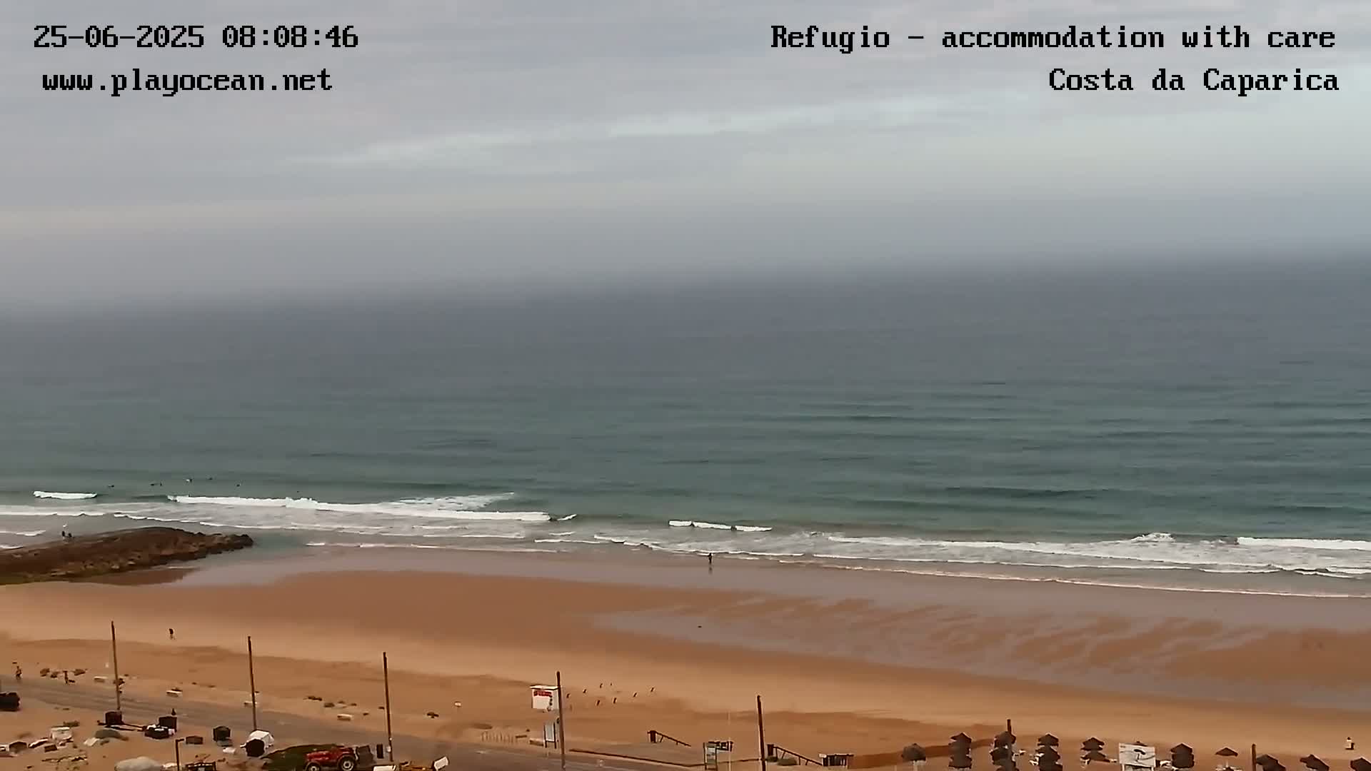 A wide shot shows a sandy beach with gentle waves lapping the shore under an overcast sky.