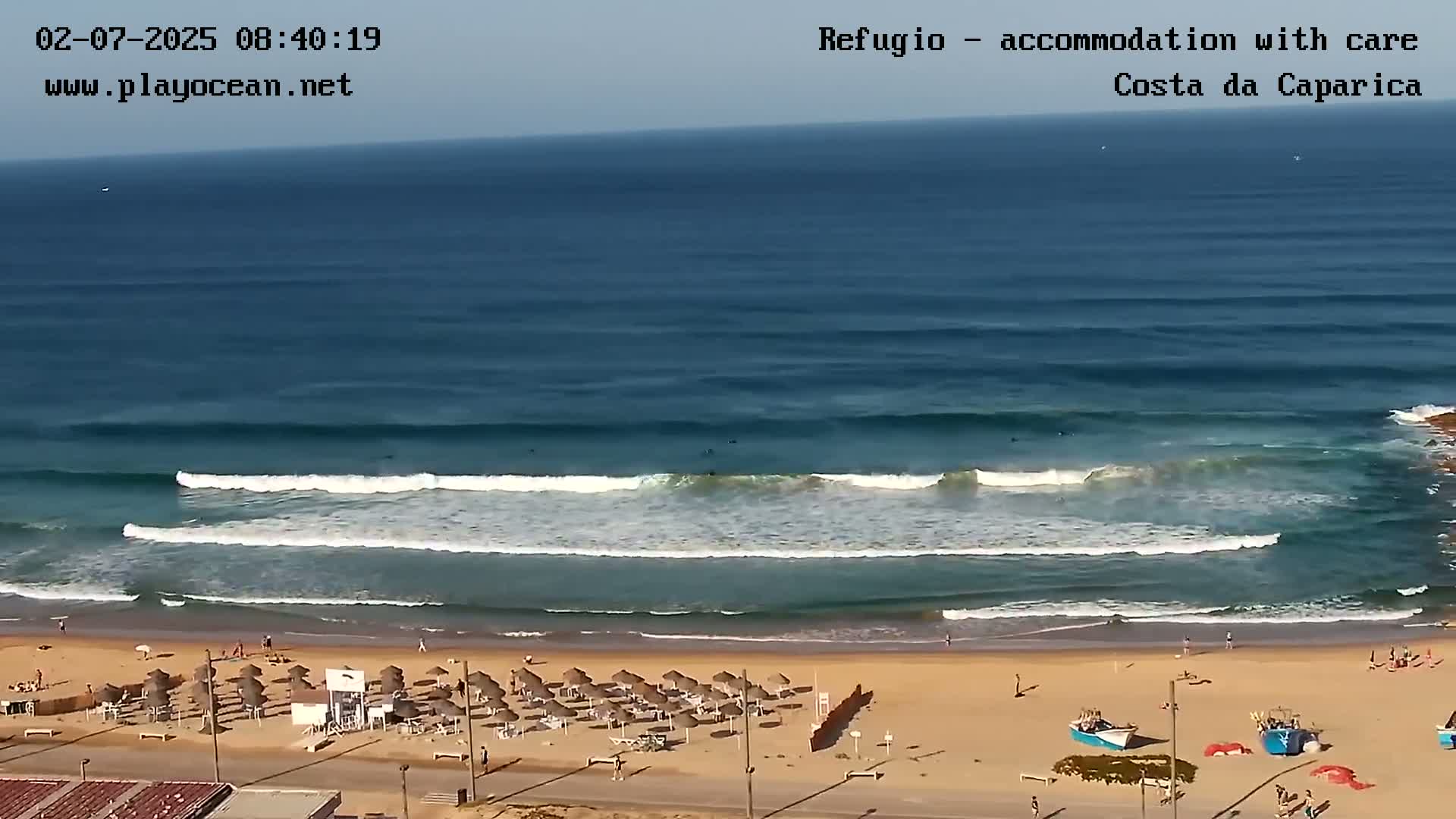 A sandy beach with rows of umbrellas and a few people is shown, with several surfers riding waves in the ocean under a clear sky.
