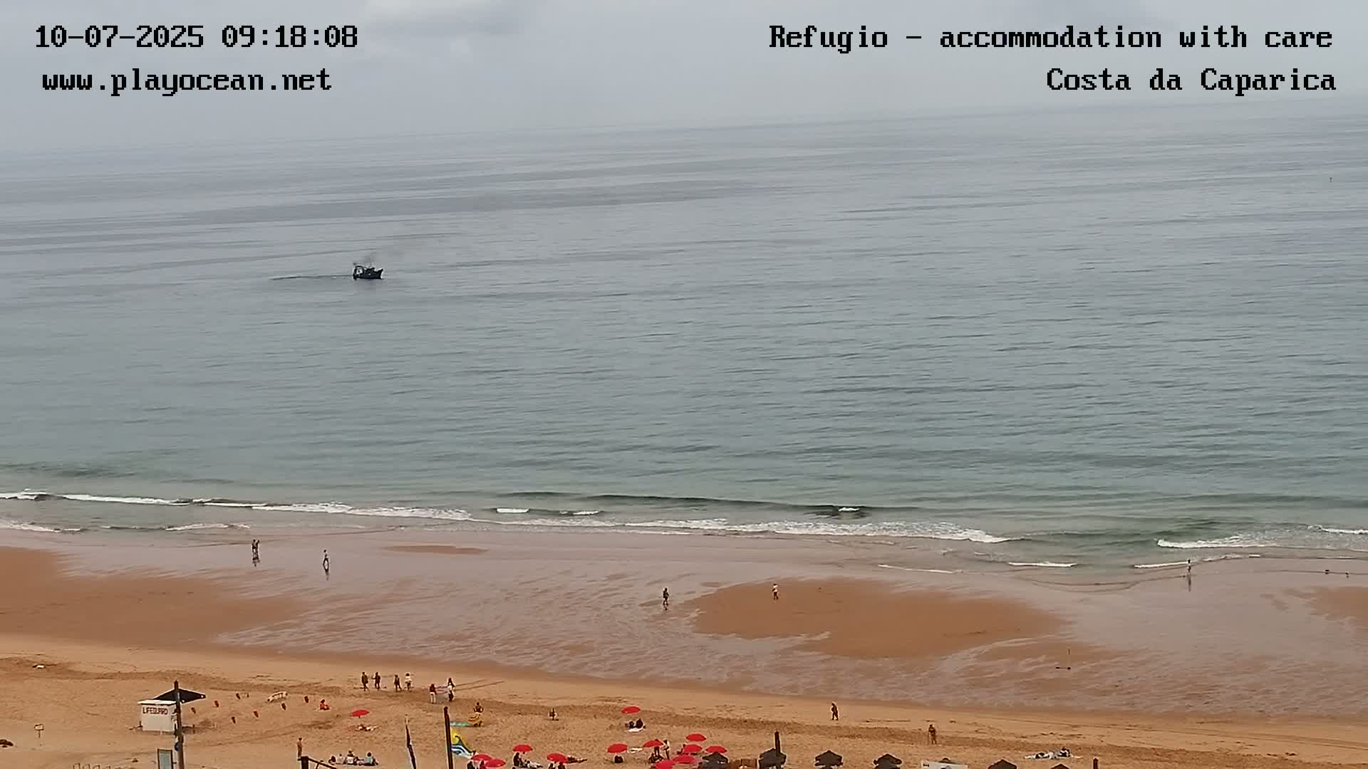 A sandy beach with a few people and red umbrellas is shown, with calm ocean waves lapping the shore under an overcast sky, and a small boat visible on the water.