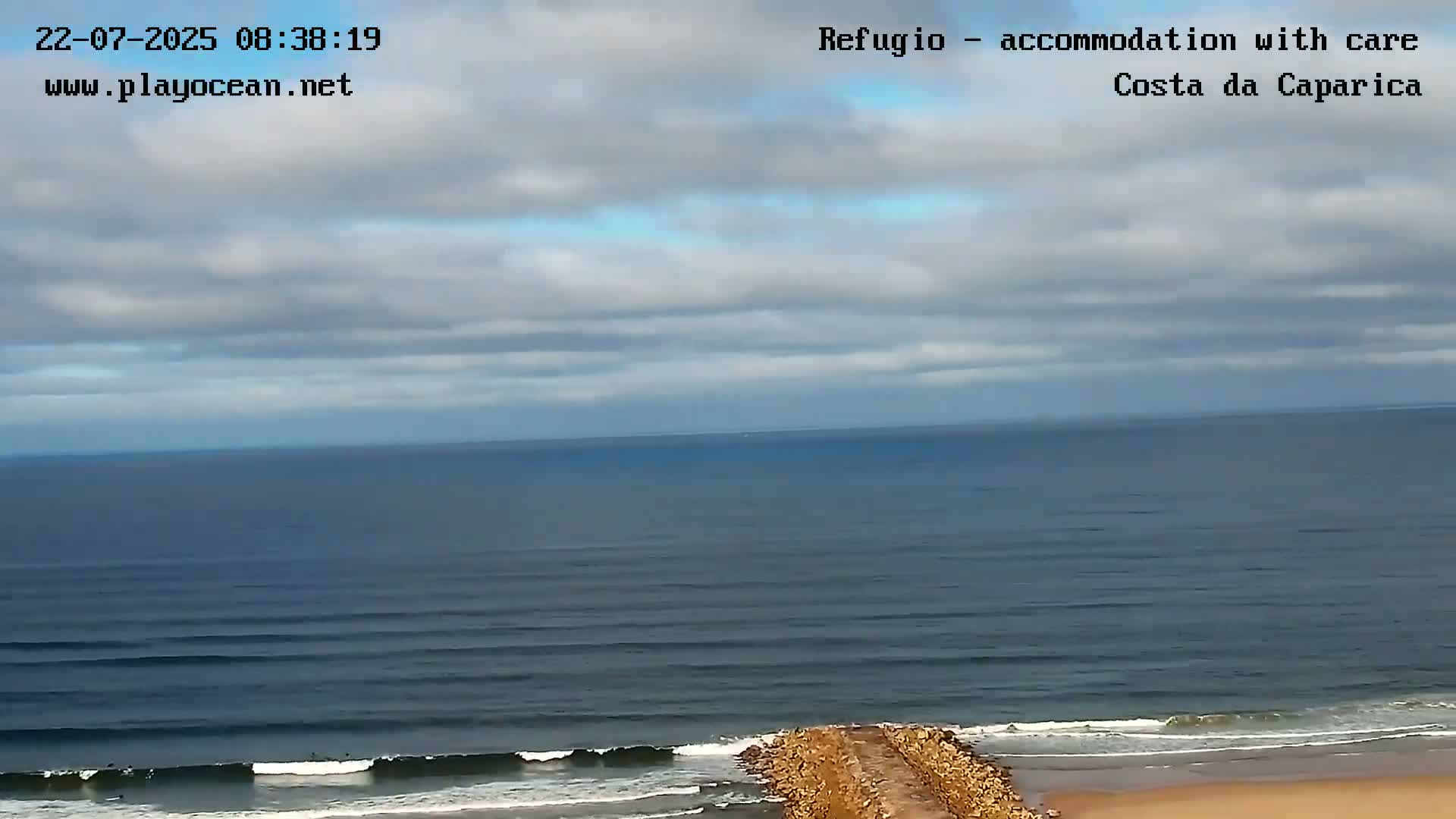An ocean scene under a partly cloudy sky shows waves breaking on a sandy shore near a rock jetty.