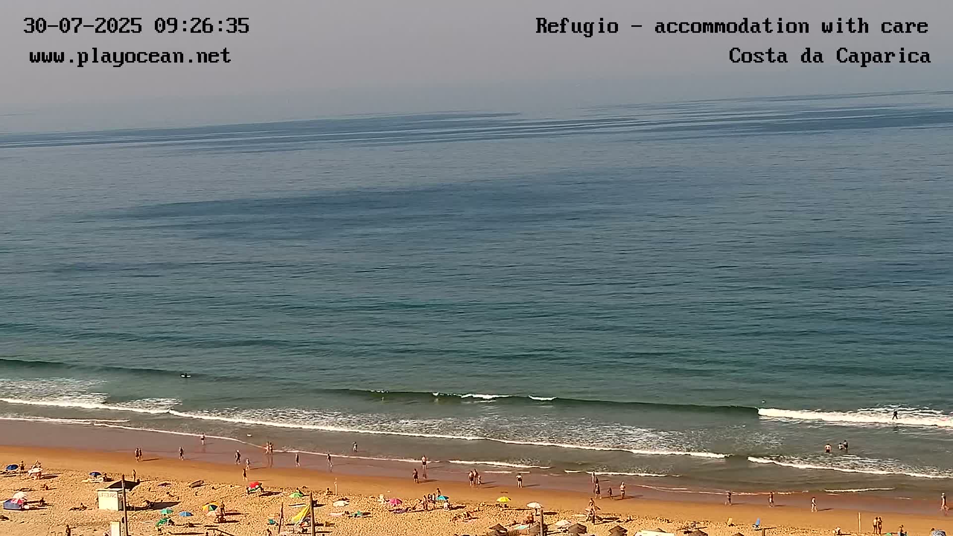 A sandy beach with people sunbathing and swimming is gently lapped by waves on a calm, clear ocean.
