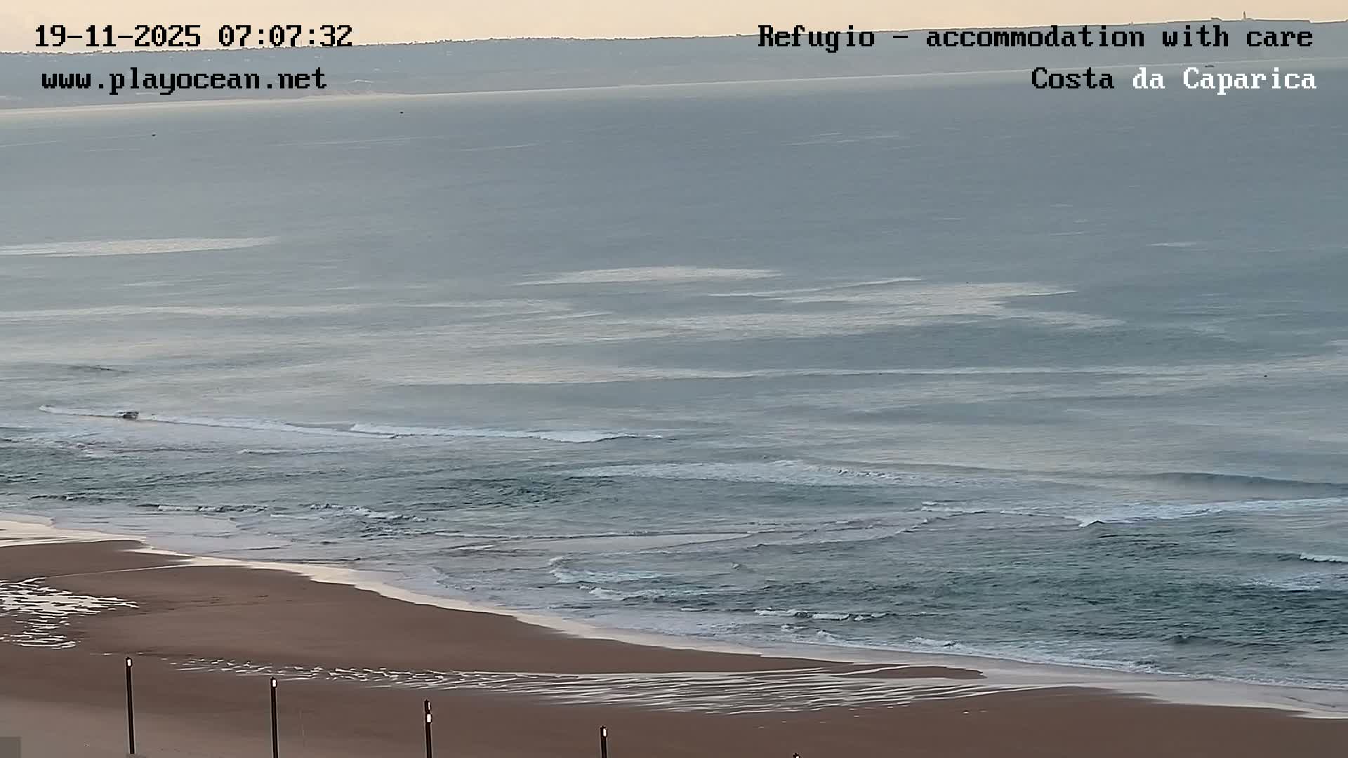 An expansive view captures a sandy beach meeting a gray-blue ocean with gentle breaking waves under a hazy early morning sky.