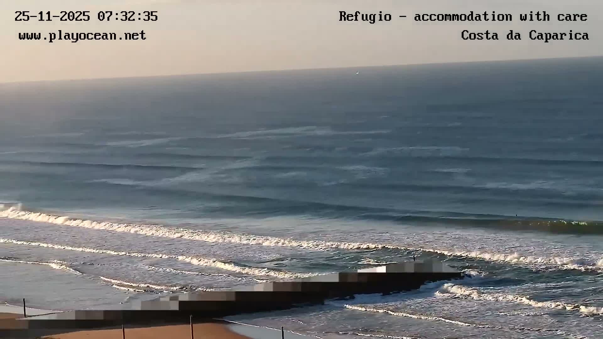 A broad view of the ocean shows multiple lines of white-capped waves approaching a sandy beach under a hazy, overcast sky.