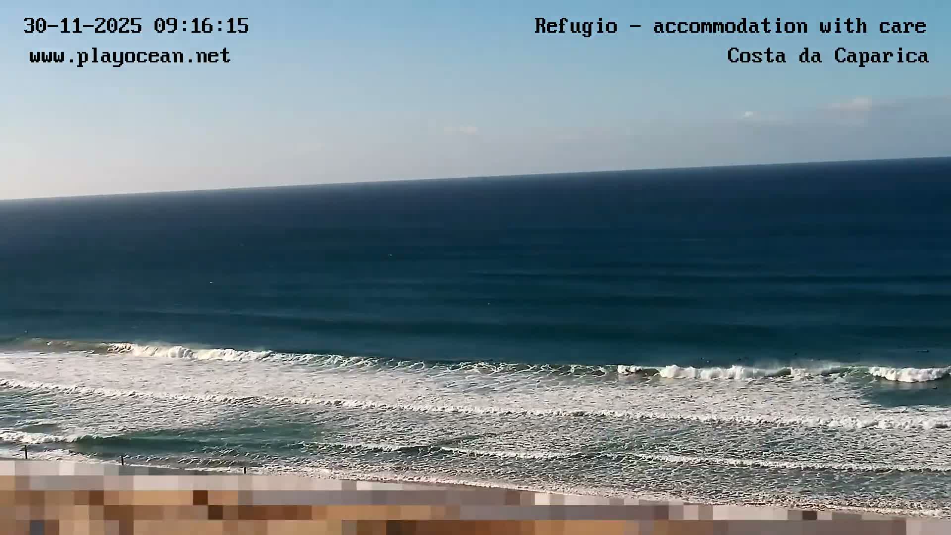 Under a clear blue sky, the vast ocean features multiple rows of white-crested waves rolling towards the distant shore, with several small dark figures visible in the water.