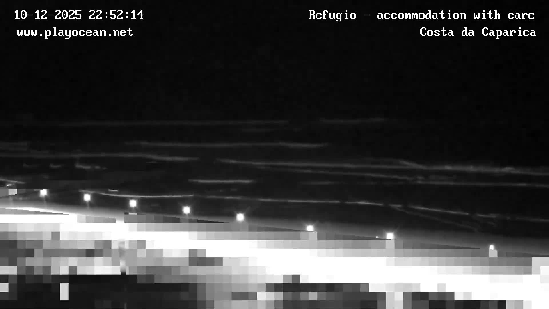 A pixelated nighttime view of an ocean beach under clear skies shows breaking waves and a row of bright lights illuminating a path or structure in the foreground.