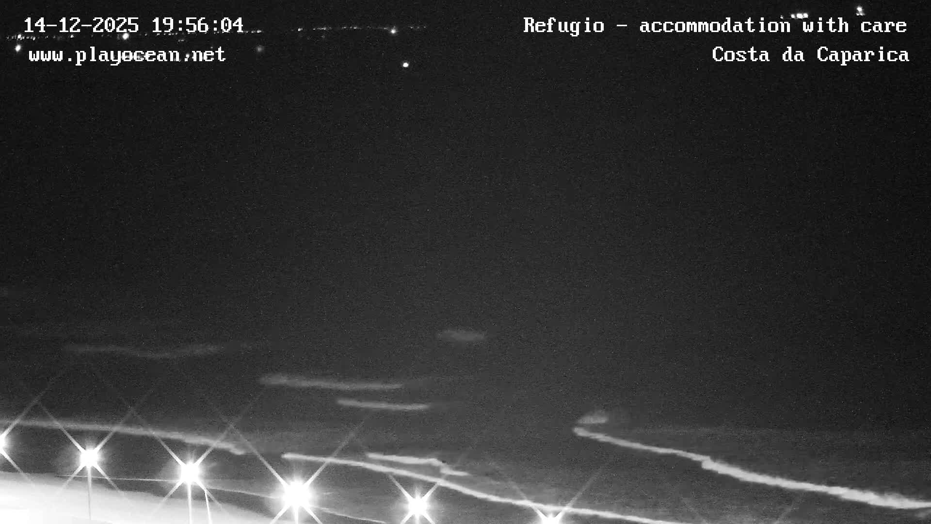 A pixelated nighttime view of an ocean beach under clear skies shows breaking waves and a row of bright lights illuminating a path or structure in the foreground.