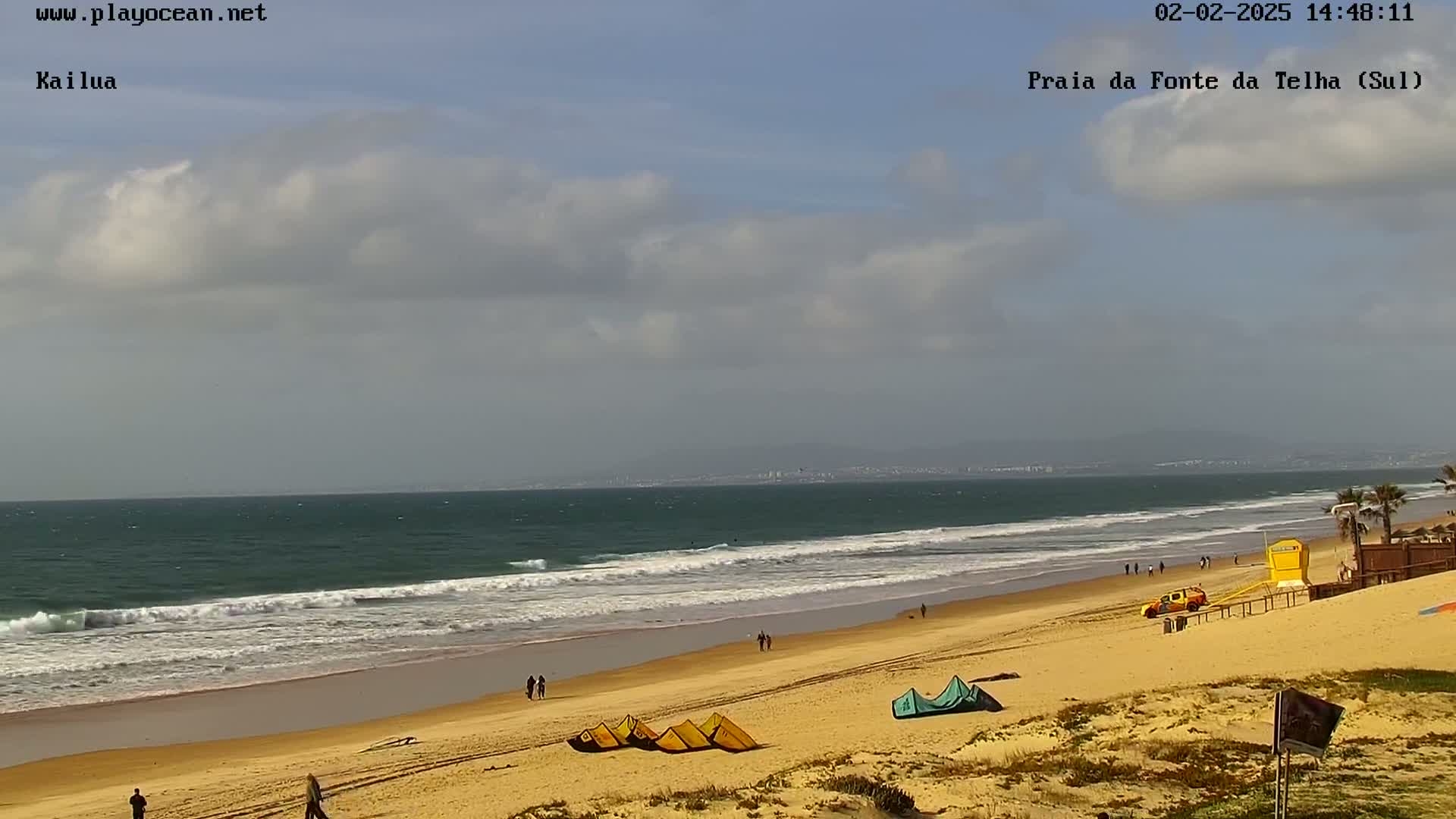 A sandy beach with a few people and some kitesurfing equipment is shown, with a partly cloudy sky and ocean waves rolling in.