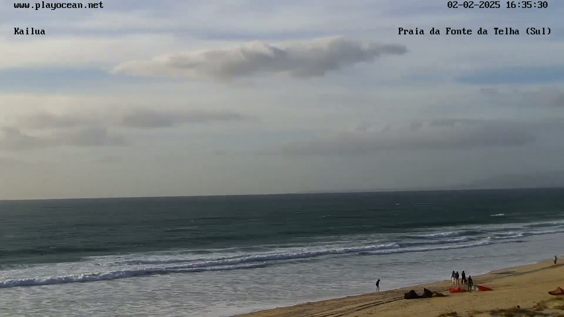 A mostly cloudy day at a beach shows a dark ocean with small waves rolling onto a sandy shore where several people are walking.