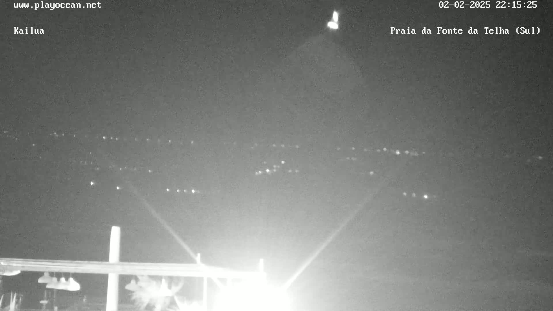 A nighttime, low-light view shows a distant line of lights, possibly from a town or city, with a brighter light source and beams in the foreground.
