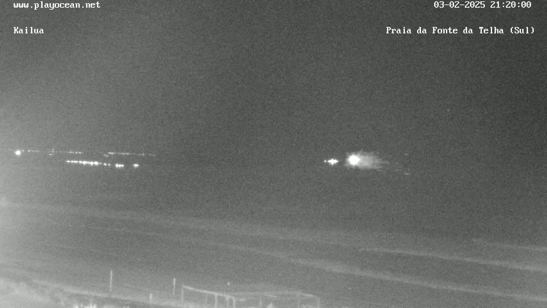 A nighttime, low-light image shows a dark beach with a distant line of lights and a bright streak of light in the sky.
