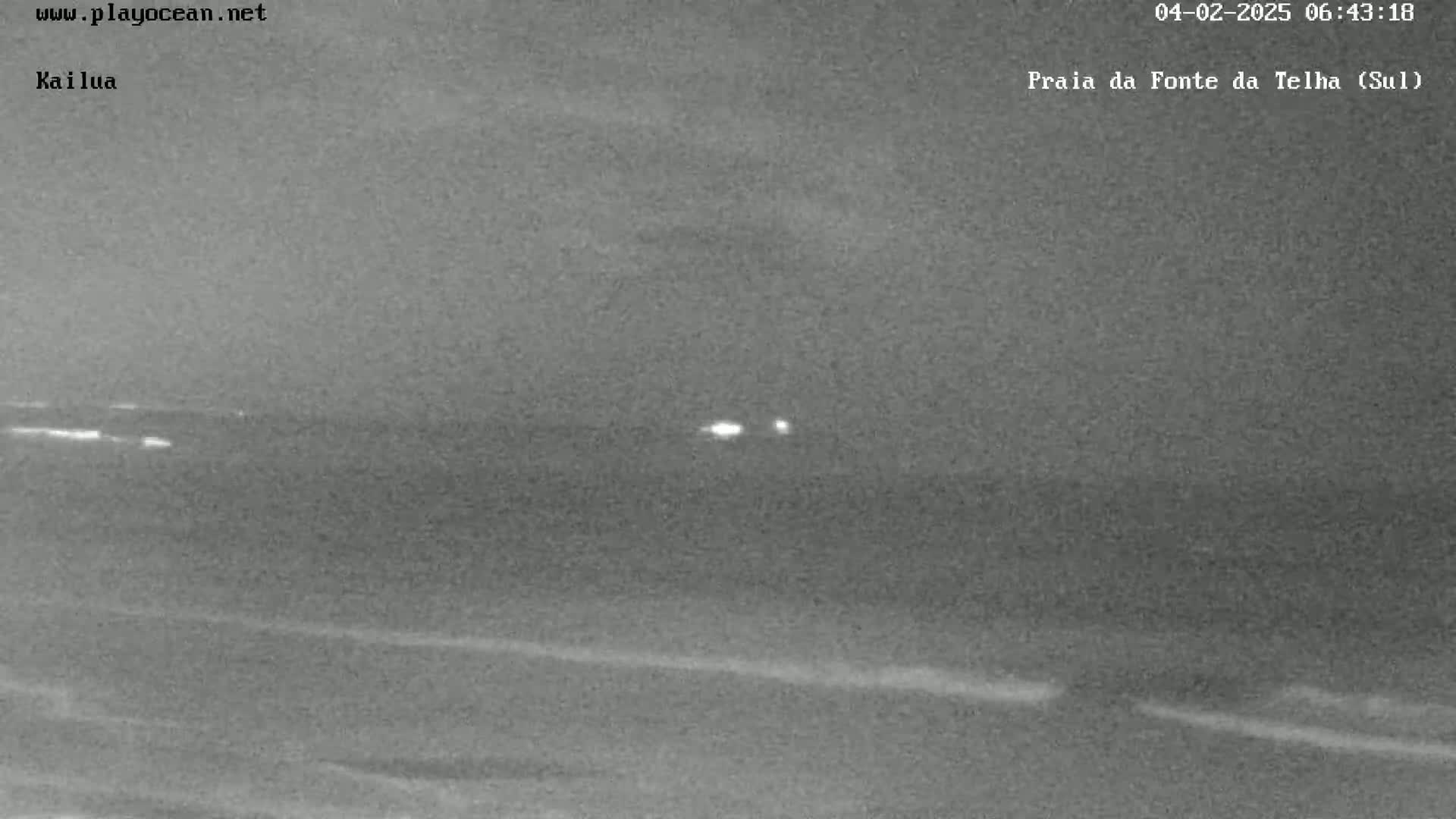 A nighttime, low-light image shows a dark beach and ocean with faint lights visible on the horizon under a cloudy sky.