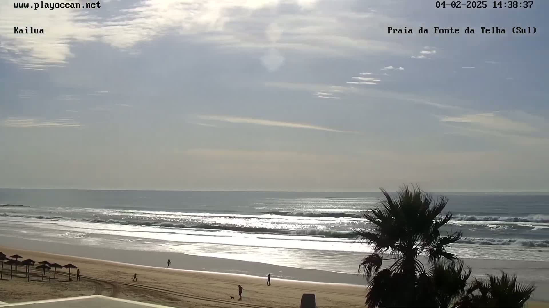 A sandy beach with gentle waves under a mostly cloudy sky, a few people are walking along the shore near some palm trees.
