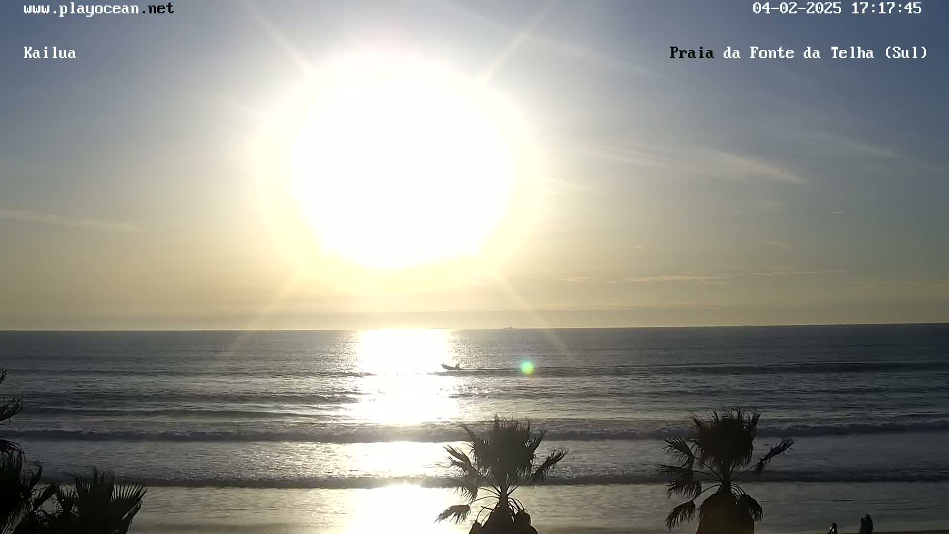 A bright sun shines over a calm ocean, with gentle waves lapping a sandy beach lined with palm trees.
