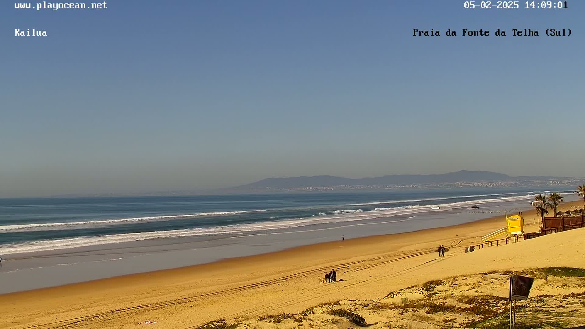 A long sandy beach with gentle waves under a clear blue sky, featuring a few people and a lifeguard stand in the distance, backed by distant mountains.