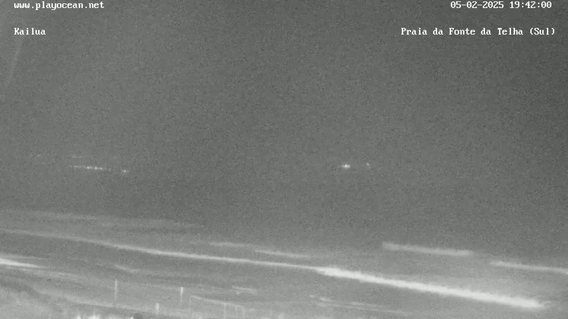 A nighttime, low-light view of a beach with gently rolling waves and distant lights visible on the horizon.
