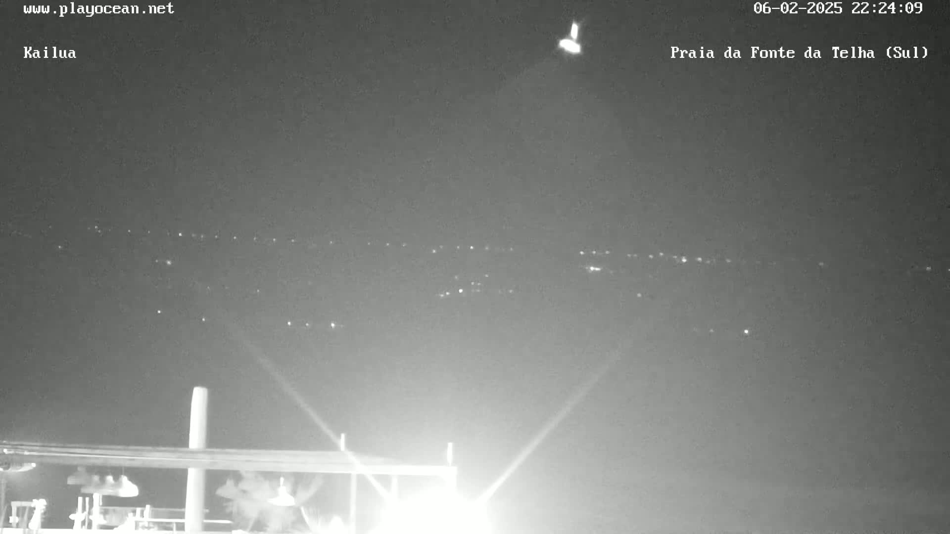 A nighttime long shot shows a distant line of lights on the horizon, a bright light source at the top center, and a dark structure in the bottom of the frame illuminated by bright lights.