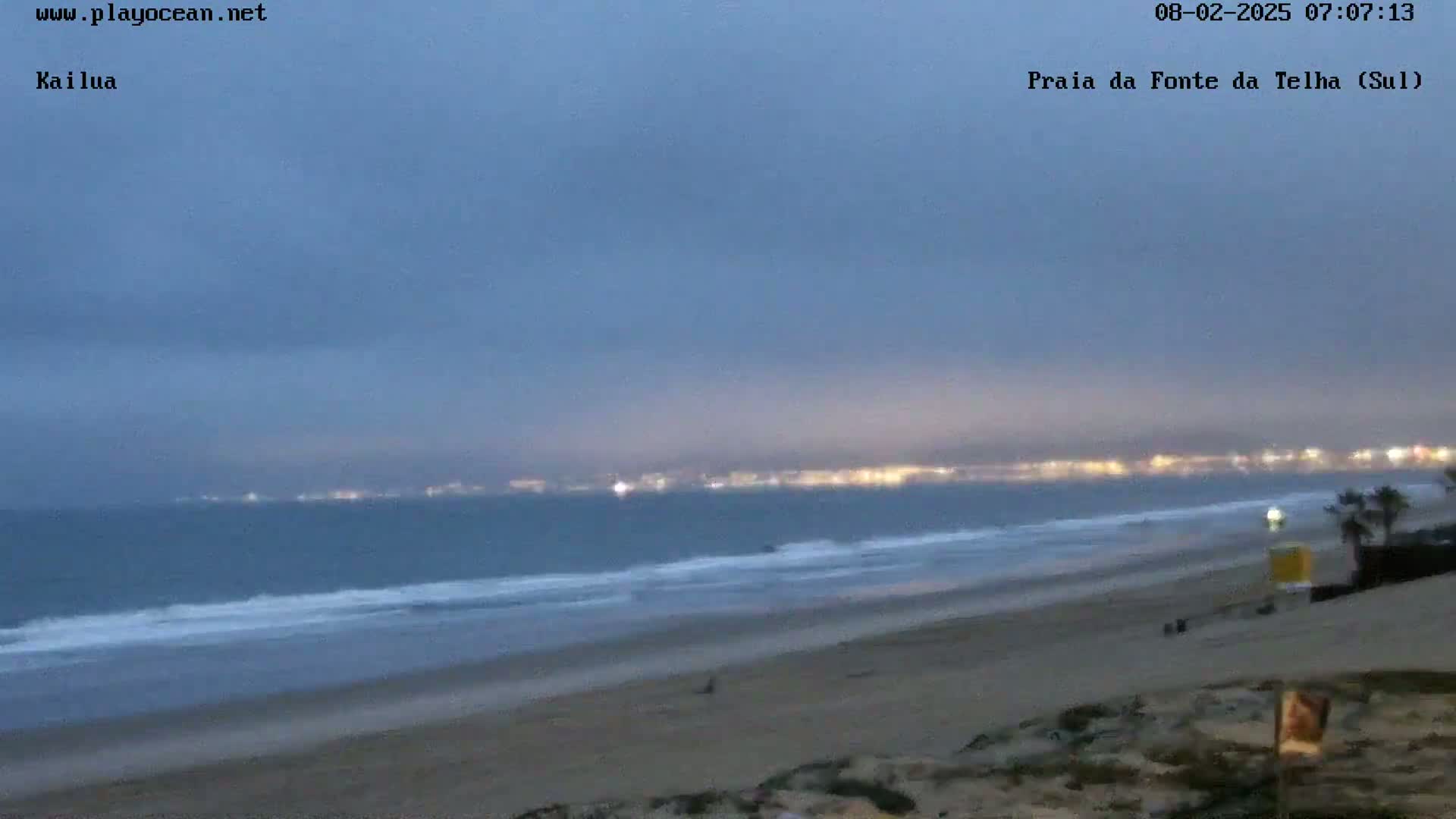 A dark, overcast sky hangs over a beach at twilight, with a distant city skyline faintly illuminated on the horizon.