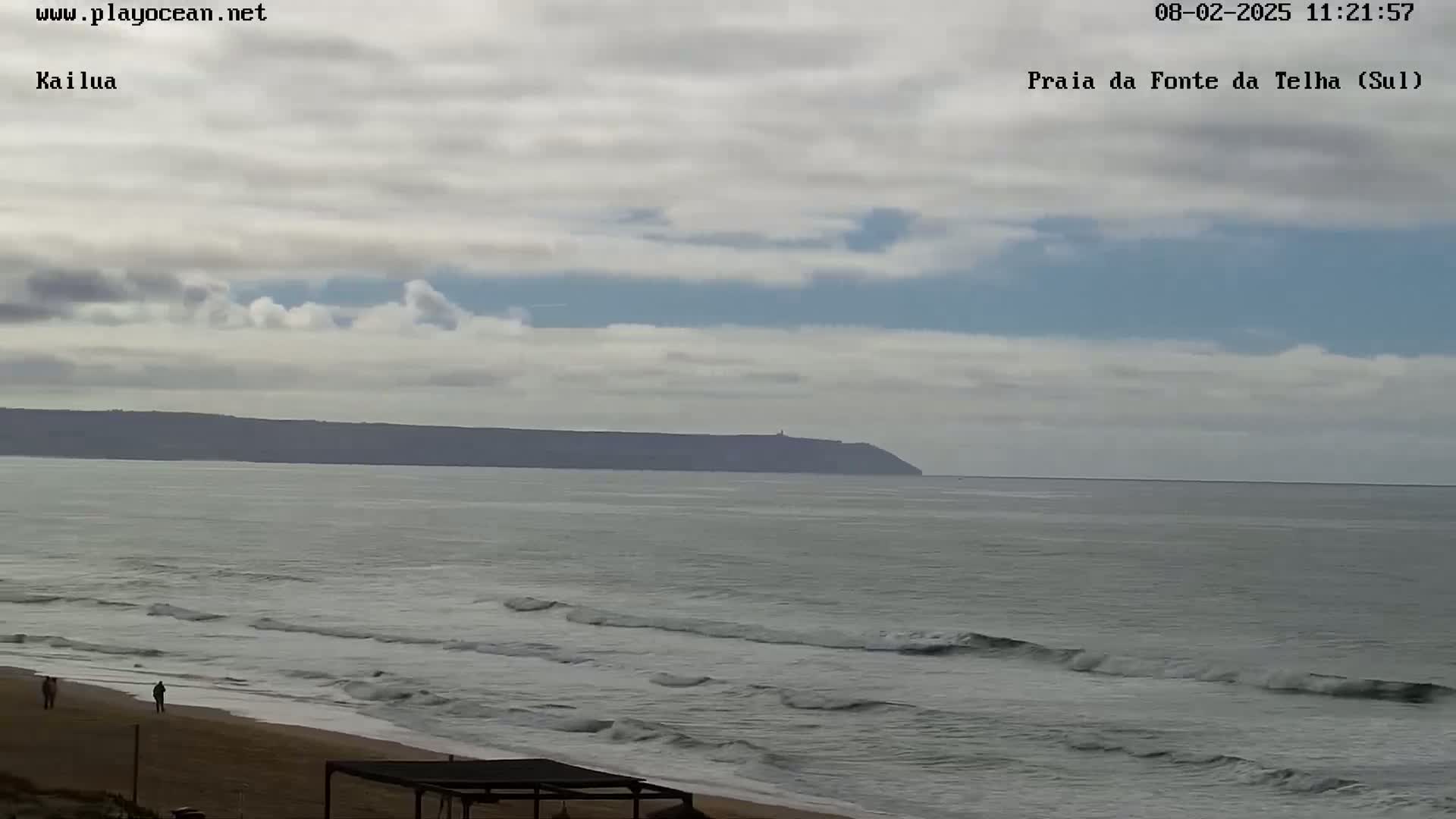 A sandy beach with gentle waves laps the shore under a mostly cloudy sky, with a distant landmass visible on the horizon.