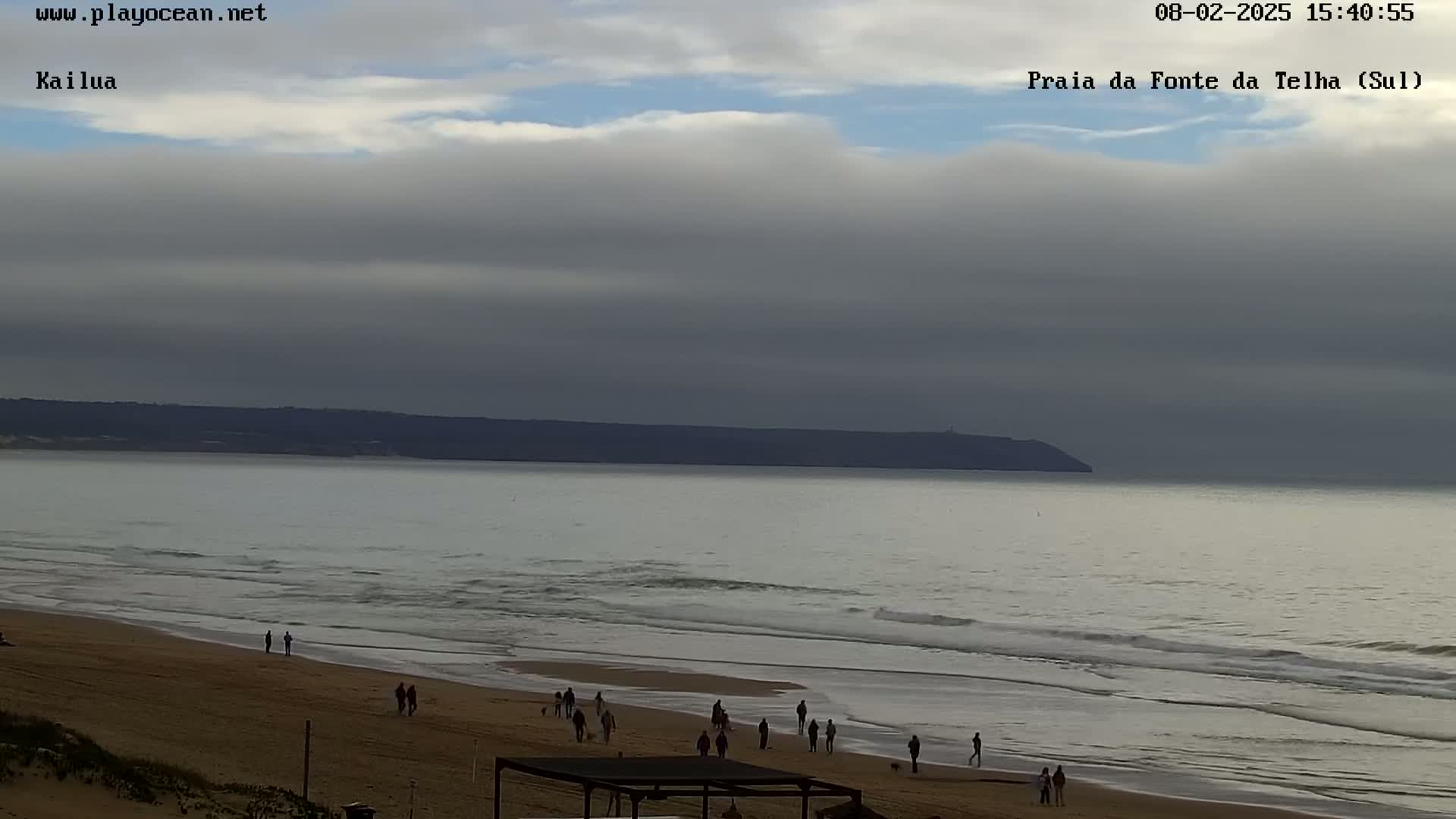A sandy beach with several people walking along the shore and in the water under a mostly cloudy sky, with calm waves rolling in from a calm ocean and a distant landmass visible on the horizon.