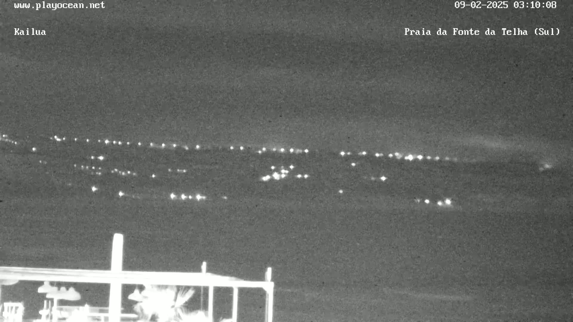 A nighttime long shot shows a distant collection of city lights under a clear, dark sky, viewed from a high vantage point near a structure.
