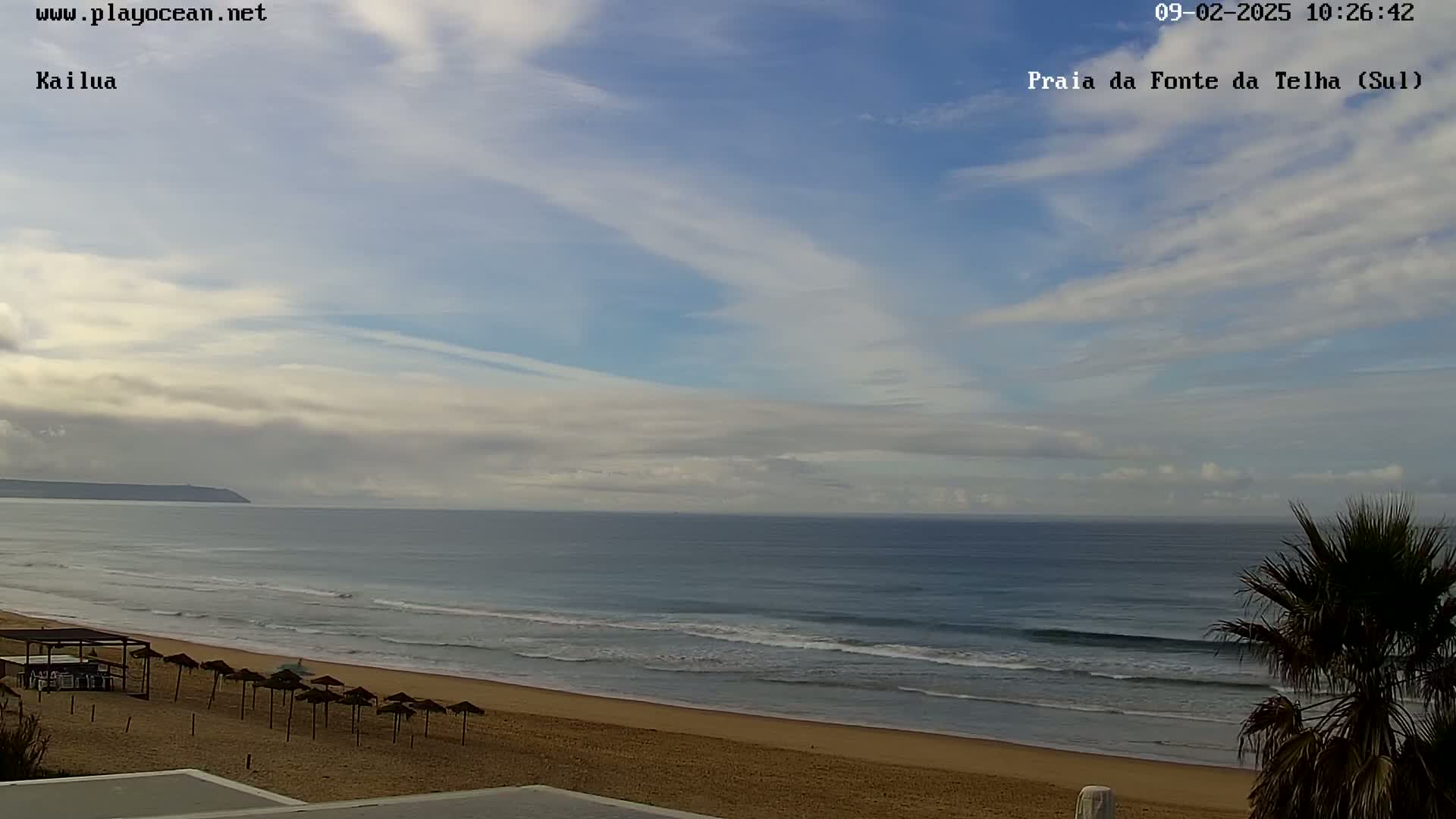 A sandy beach with calm ocean waves stretches beneath a mostly clear sky with thin, wispy clouds.