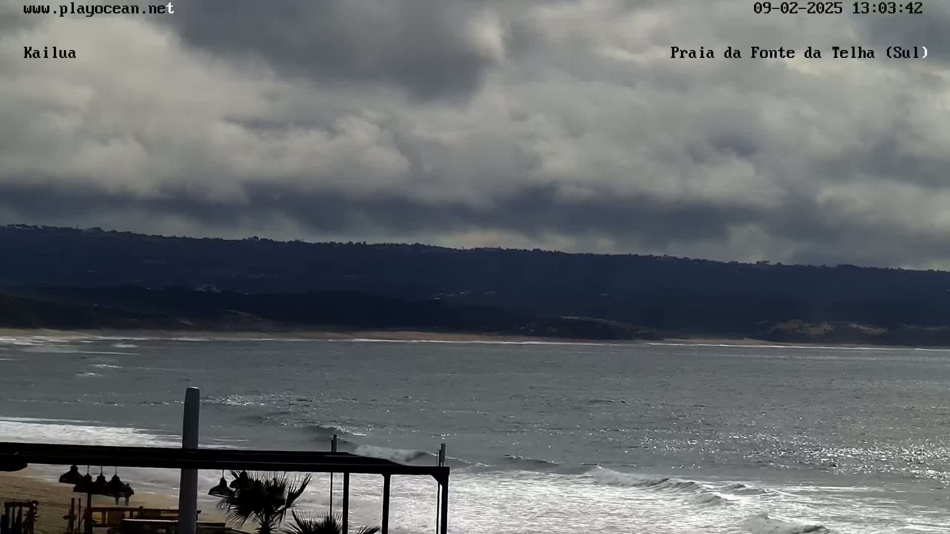 A mostly cloudy day shows a beach with gentle waves lapping the shore, a dark mountain range in the background, and a partially visible outdoor structure in the foreground.