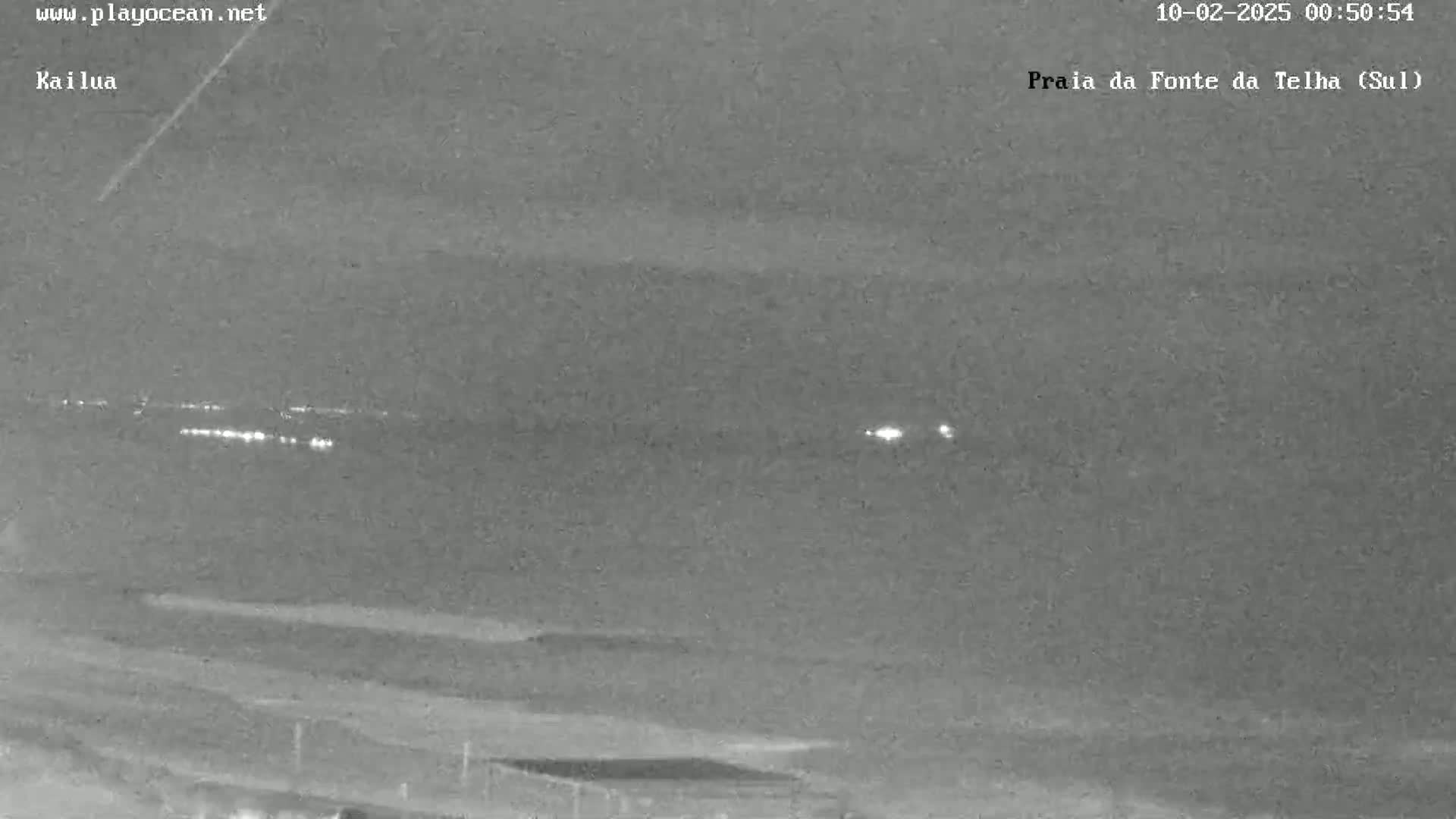 A nighttime, low-light view shows a dark beach and ocean with a line of distant lights on the horizon.