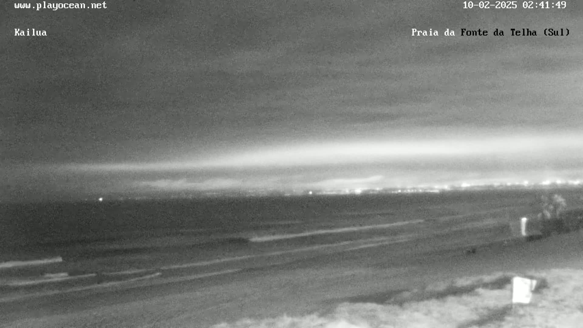 A nighttime view of a dark ocean beach with a city skyline faintly visible in the distance under a mostly clear sky.