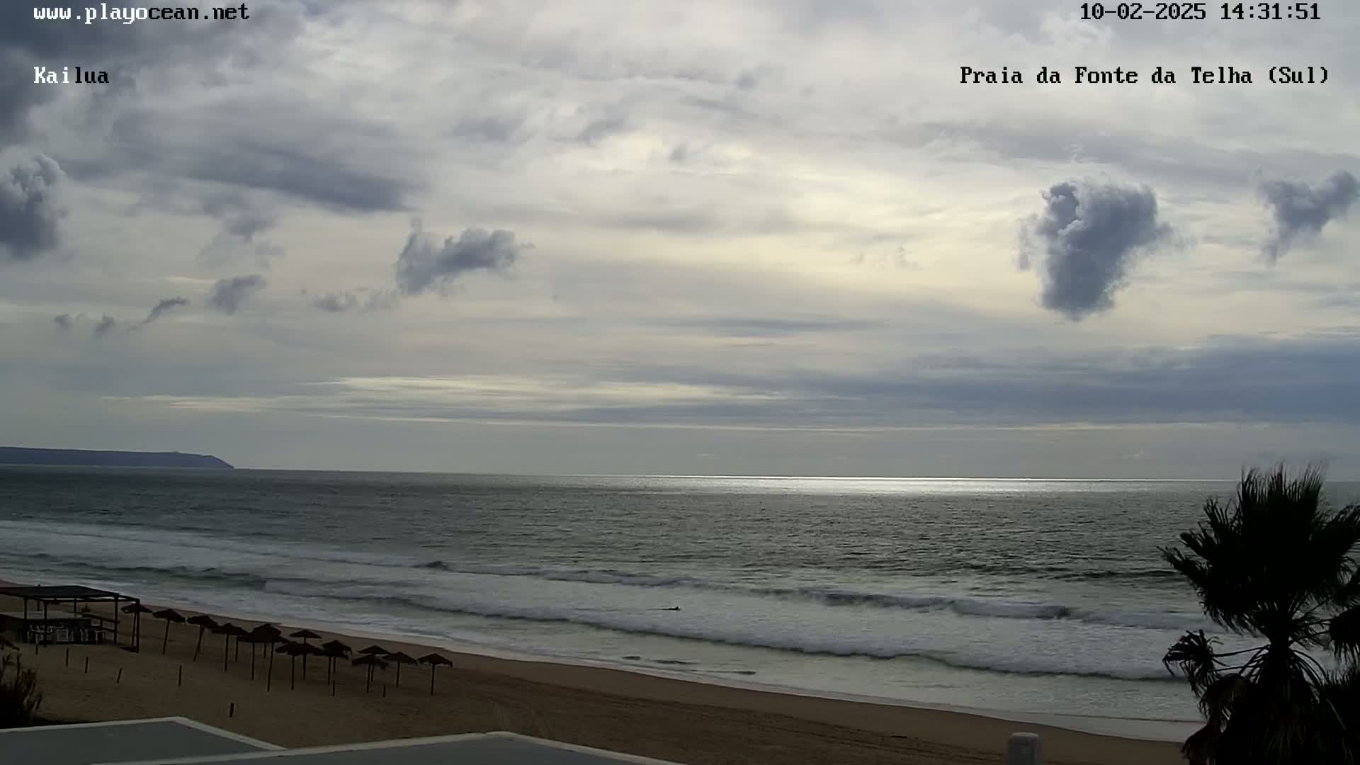 A mostly cloudy sky overlooks a sandy beach with calm waves and a few closed umbrellas under a mostly cloudy sky.