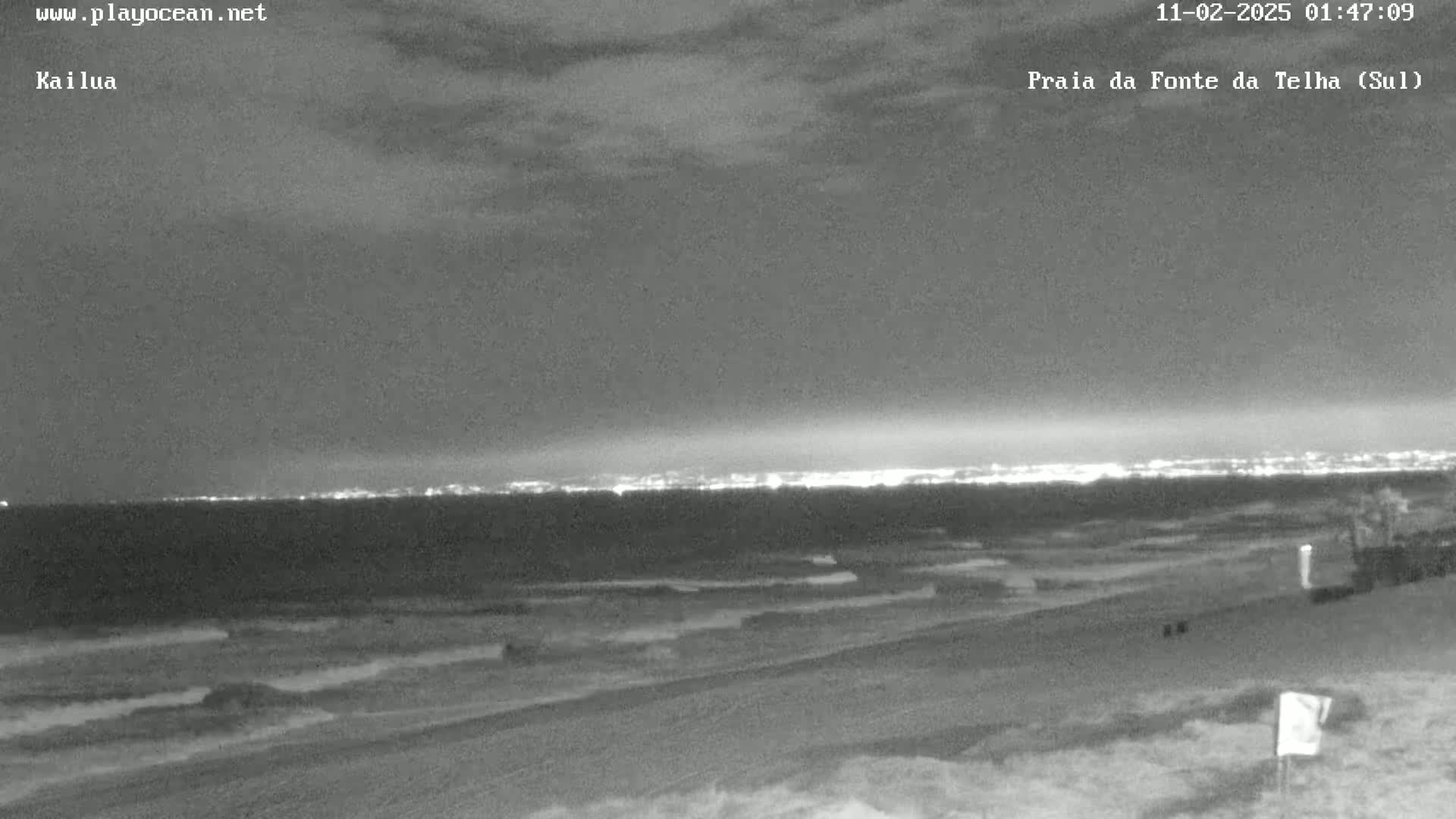 A nighttime beach scene shows dark, gently rolling waves lapping onto a sandy shore, with city lights visible on the horizon under a mostly cloudy sky.