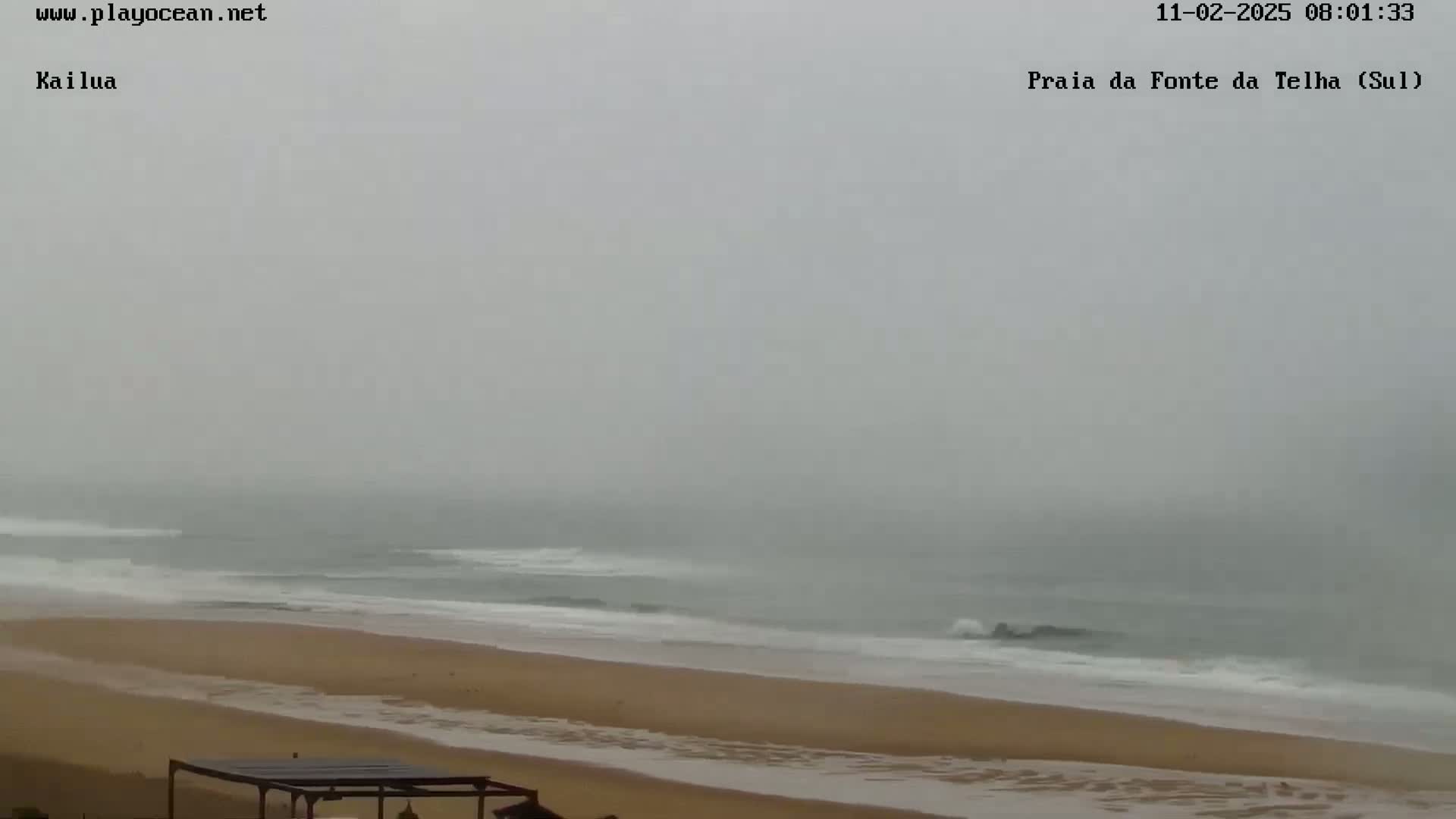 A foggy beach scene shows a sandy shore and ocean with small waves under an overcast sky.